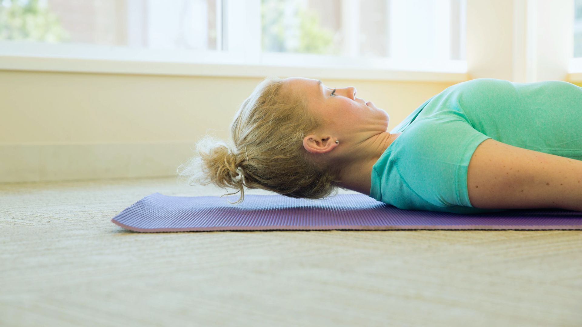 A person with blonde hair lies on a purple yoga mat, performing a neck relaxation exercise. They are indoors, wearing a turquoise shirt, and appear calm.