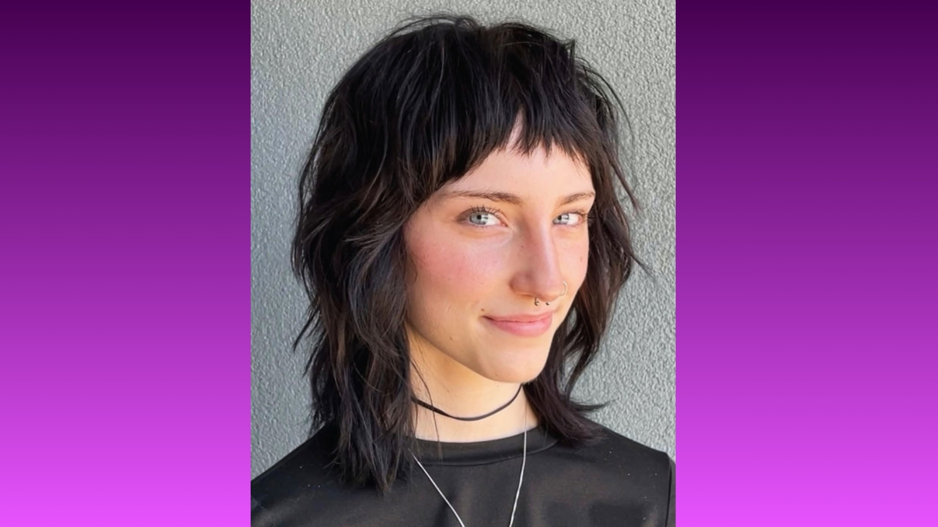 Person with short, textured dark hair and a septum piercing smiles slightly against a gray background. The image conveys a confident, edgy style.
