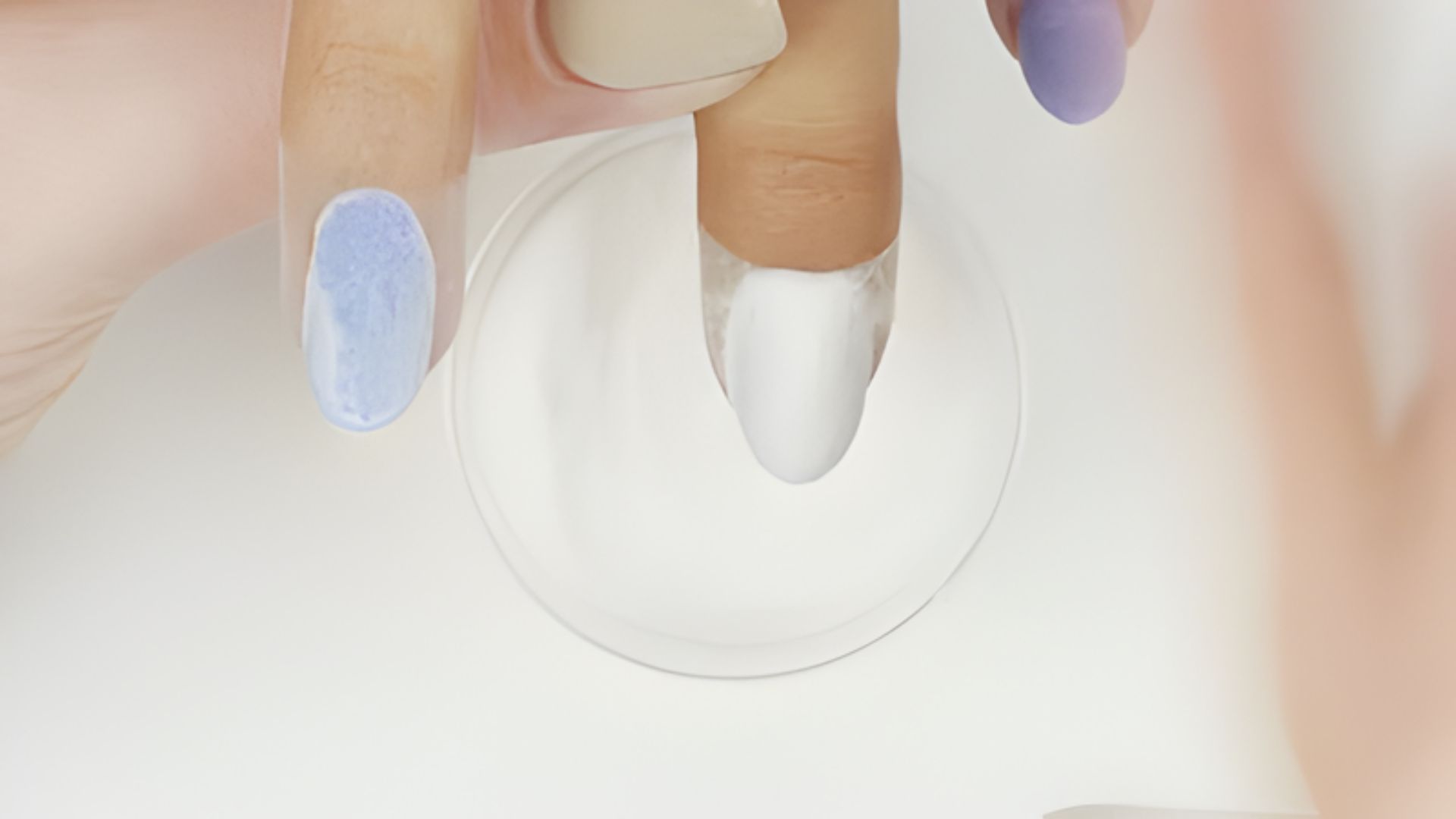 Close-up of a hand with three fingers partially visible, two nails painted in pastel blue and beige, one dipped in white powder. The scene conveys a calm, careful, and focused atmosphere typical of nail art application.