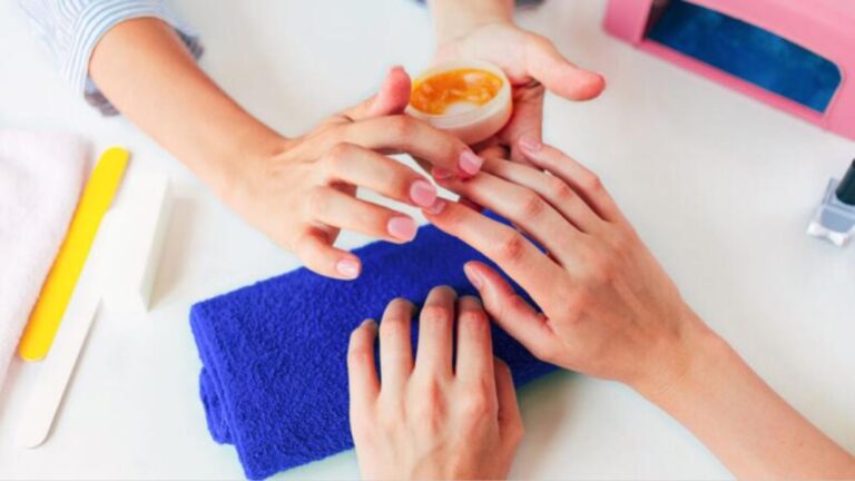 Two hands receiving a manicure at a salon, with a focus on applying cuticle cream. A blue towel and manicure tools are visible, conveying relaxation.
