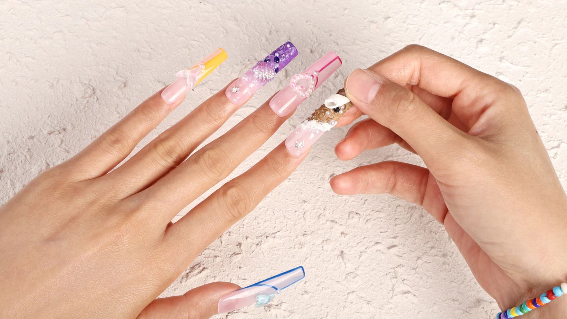 A person holds one hand steady as they apply decorative charms to long, colorful, acrylic nails on the other hand against a textured backdrop.