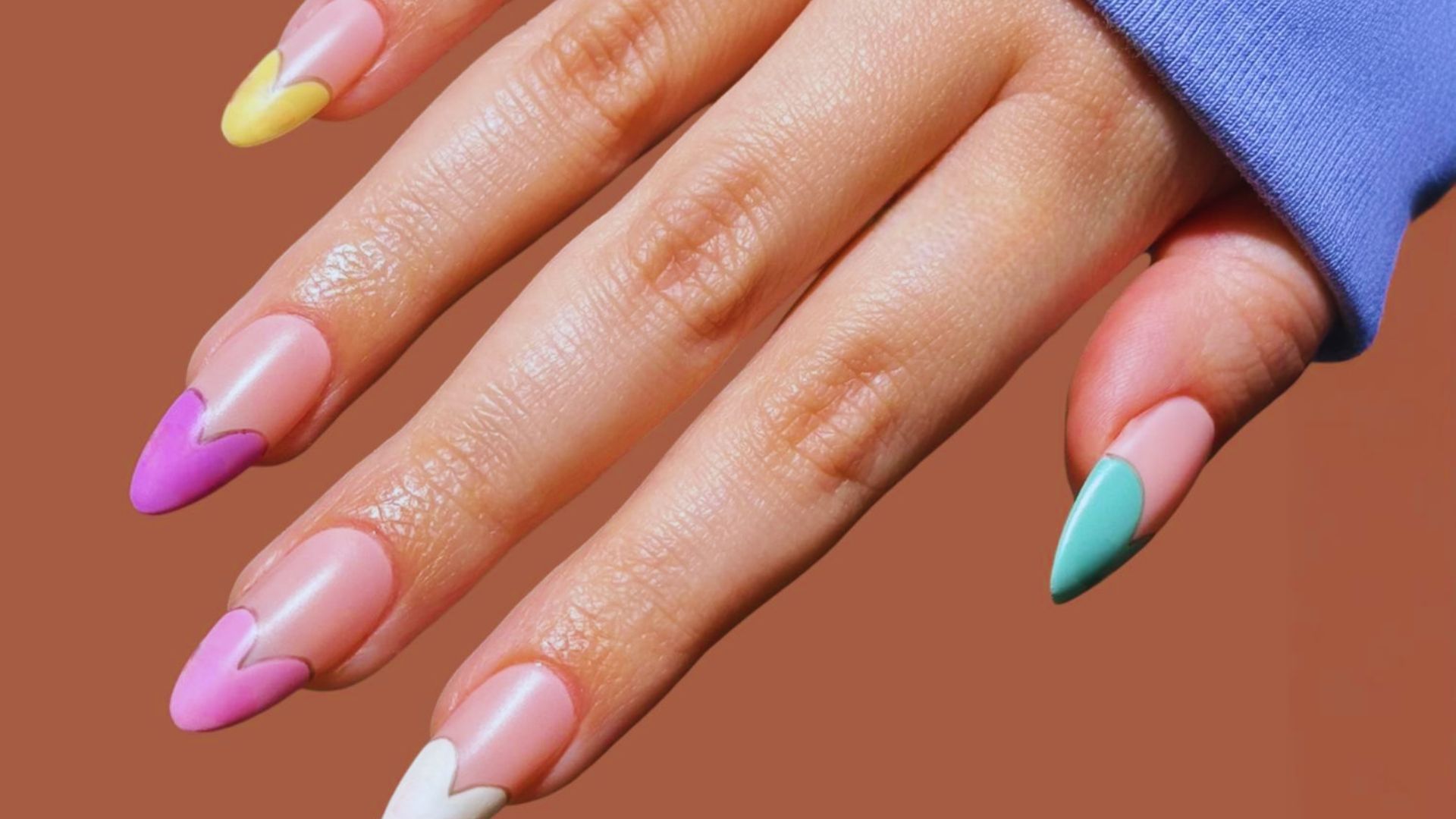 A hand with colorful, heart-shaped tips on each fingernail in shades of pink, yellow, white, and teal, against a brown background. The tone is playful and artistic.