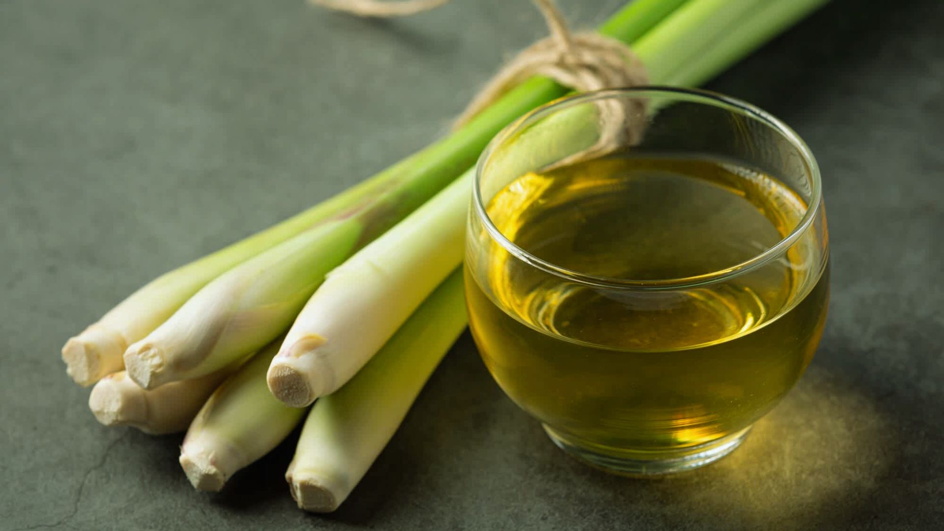 A glass cup of lemongrass tea sits on a dark surface, beside fresh lemongrass stalks. The scene conveys a sense of natural wellness and calm.