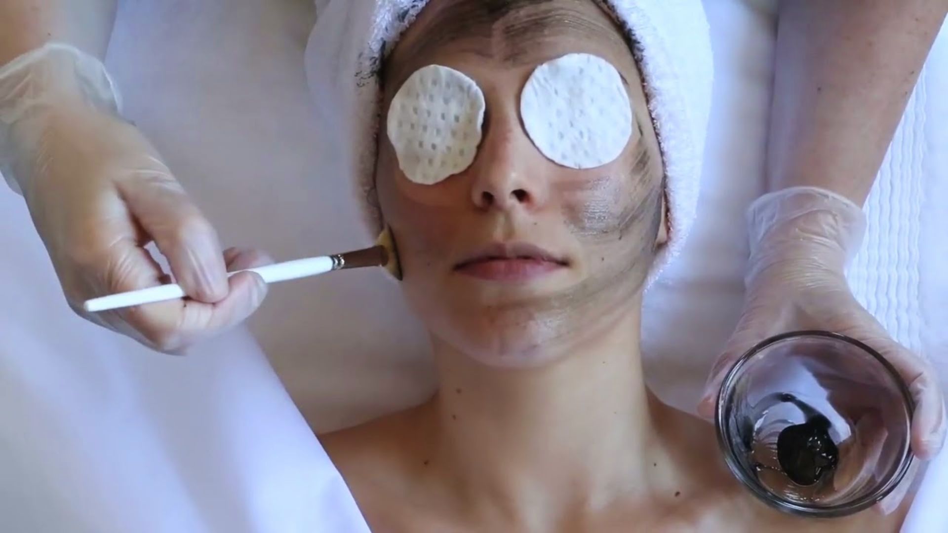 A person receiving a facial treatment lies with cotton pads on their eyes, a towel on their head. A brush applies mask, conveying calm and relaxation.