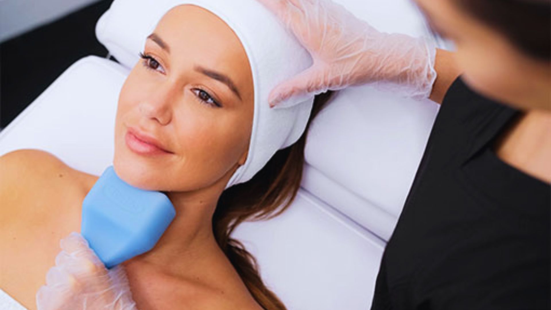 A woman lies relaxed on a spa bed, receiving a facial treatment. A gloved professional uses a blue device on her chin. The atmosphere is calm and serene.