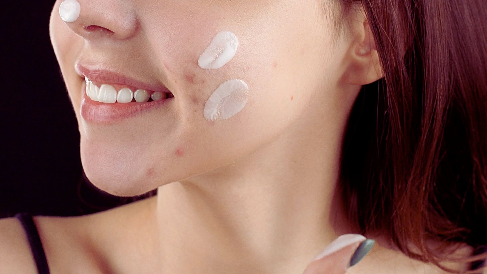 Smiling woman applies skincare cream on her cheek and nose. She has a natural complexion and wears a black top. The mood is fresh and caring.