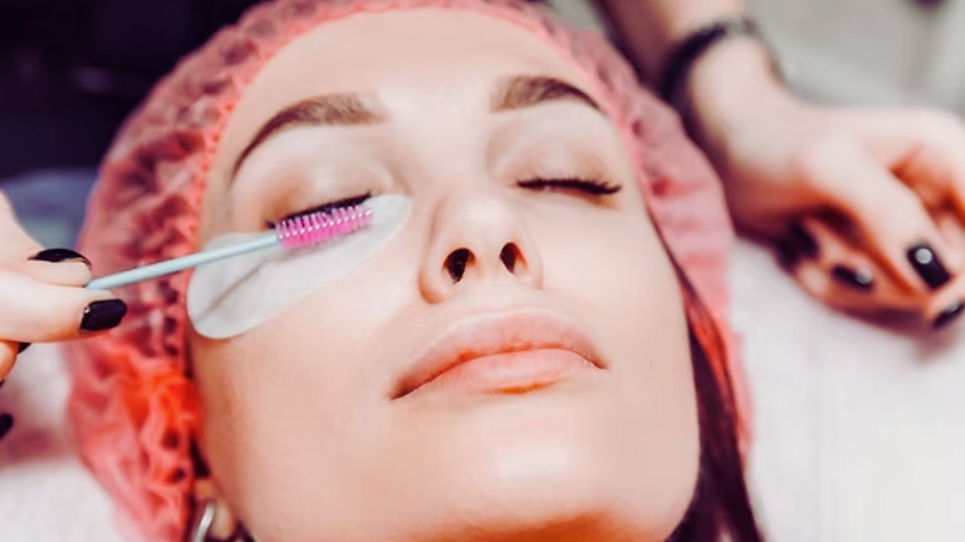 A person with a relaxed expression receives a lash treatment. Eye pads protect the skin, and a pink brush is used on the eyelashes. The tone is calm and soothing.
