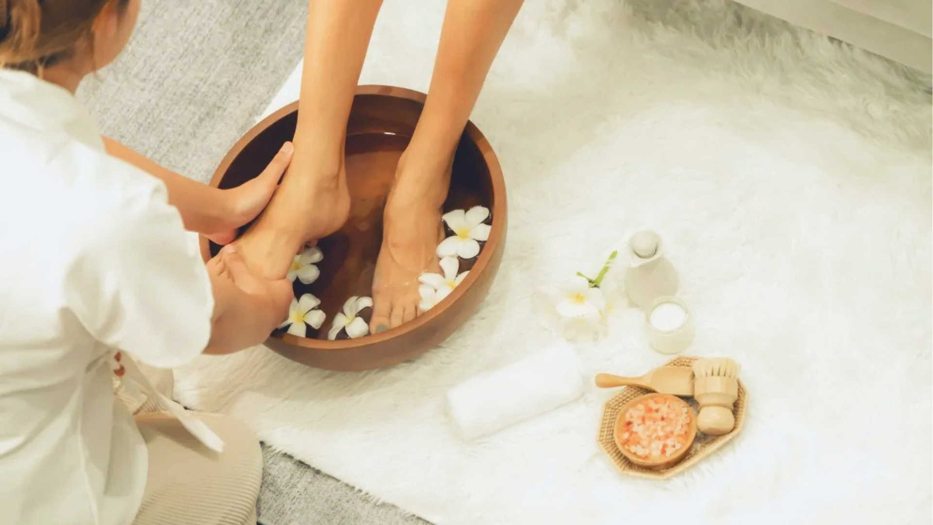 A person receives a foot soak in a wooden bowl with floating flowers, surrounded by spa items on a soft, white rug. The setting is serene and relaxing.