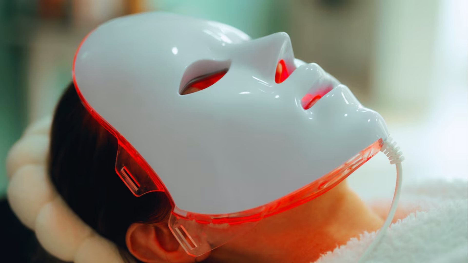 A person is wearing a LED light therapy mask, glowing red, while lying down. The setting is serene, suggesting a spa or skincare treatment.
