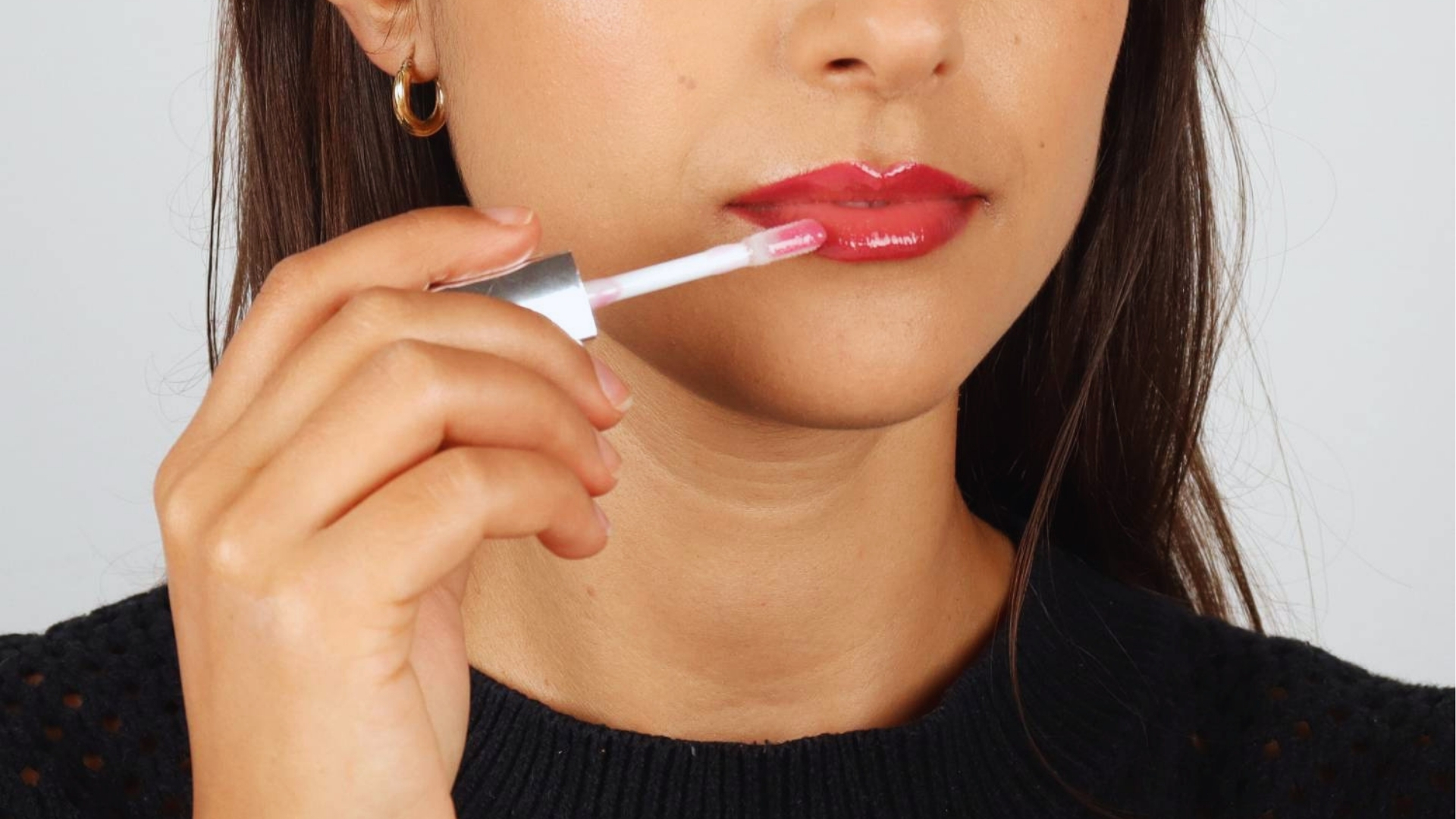 Woman applying glossy pink lip gloss to her lips, holding the applicator with her right hand. She has long brown hair and wears a black sweater.