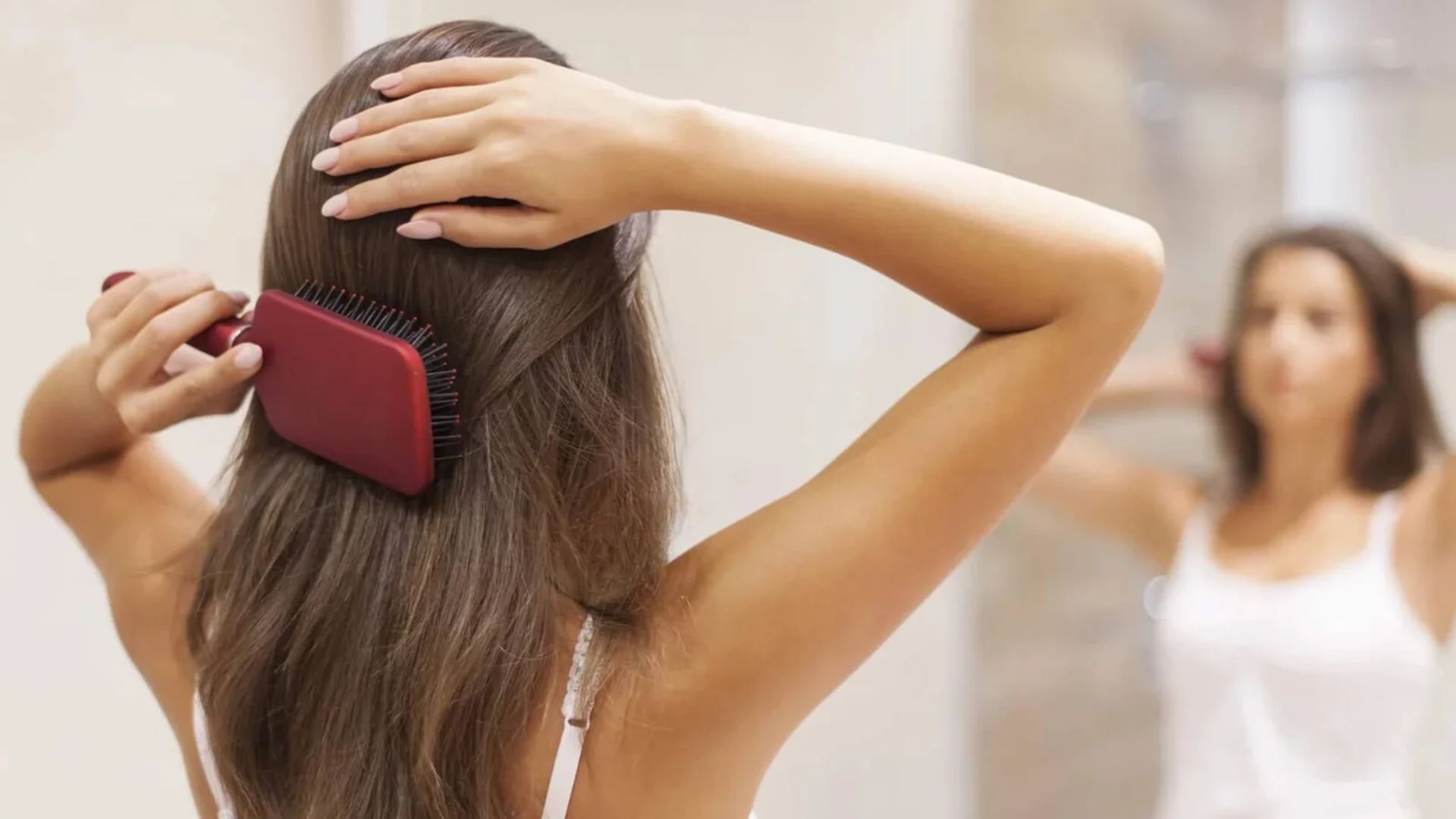 A woman is brushing her long brown hair with a red paddle brush, facing a mirror. She wears a white tank top, creating a calm, everyday routine vibe.