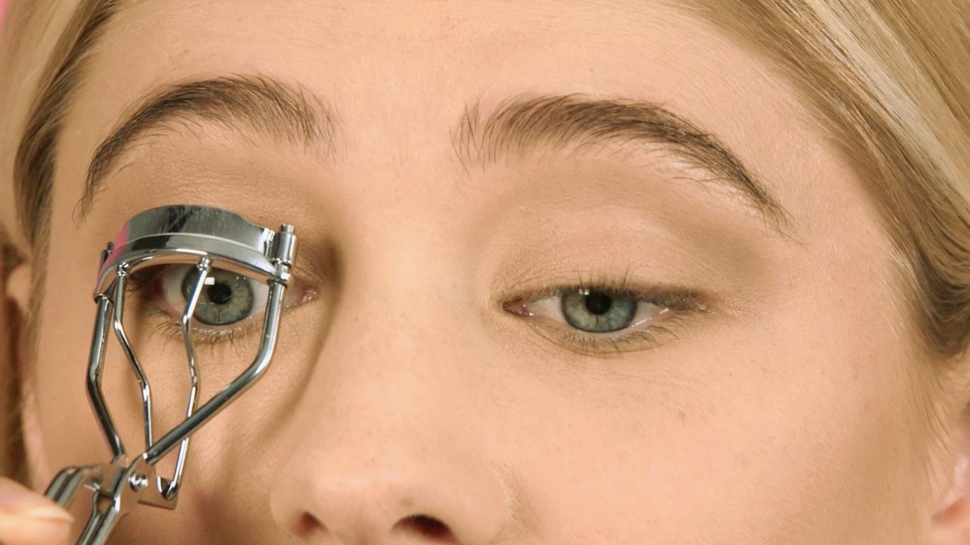 A close-up of a person using a silver eyelash curler on their right eye. The eyelids have a natural eyeshadow, and the person appears focused.