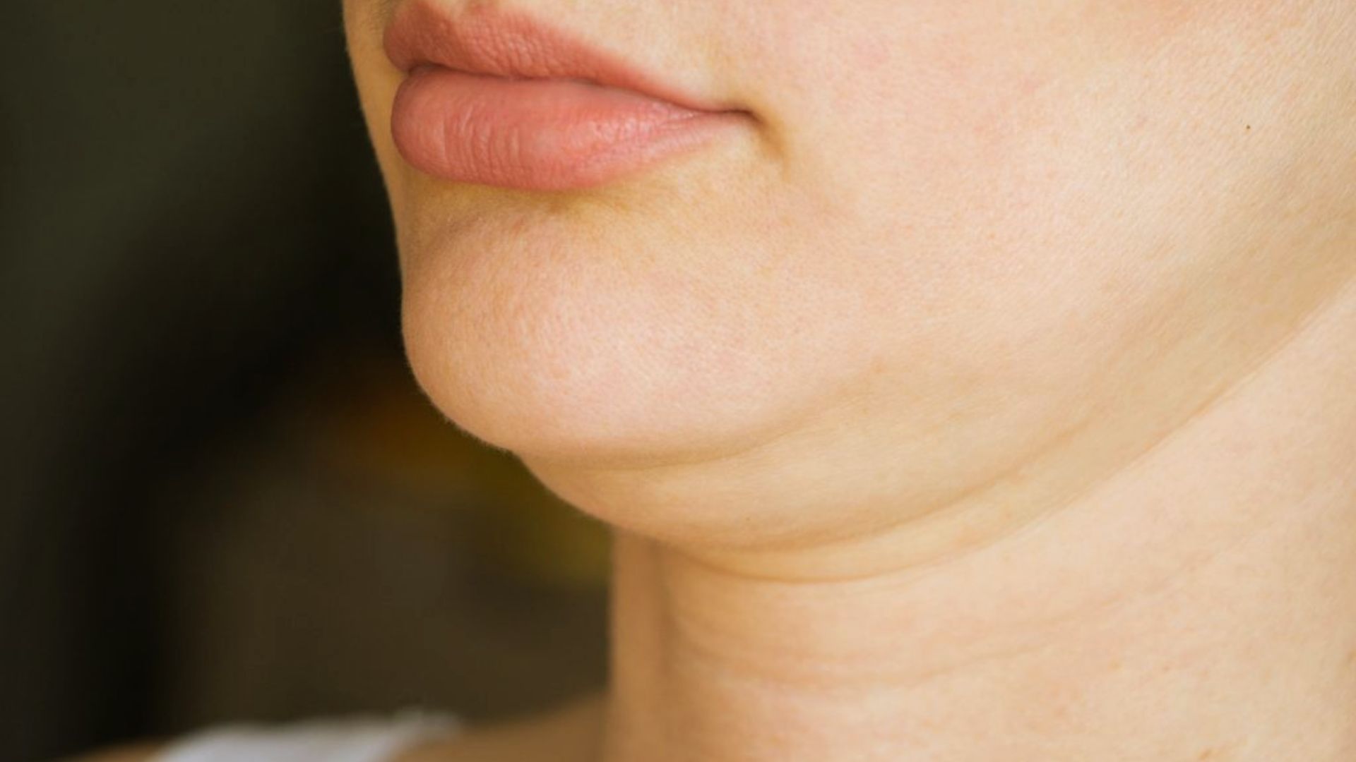 Close-up of a person's chin and neck, showing light skin with a prominent double chin. The lips are slightly parted, and the background is out of focus.
