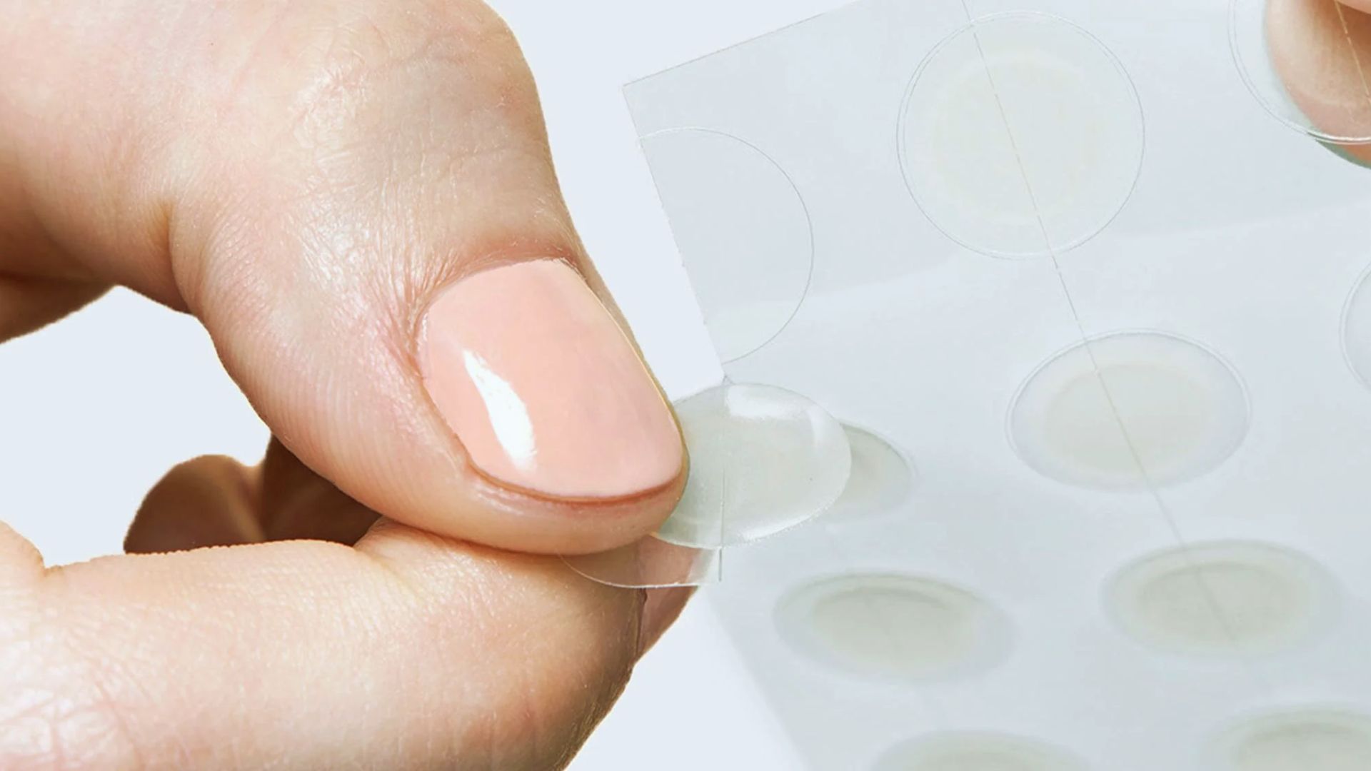 Close-up of a hand peeling a translucent pimple patch from a sheet with smooth, pale pink fingernail polish. The tone is clean and focused on skincare.