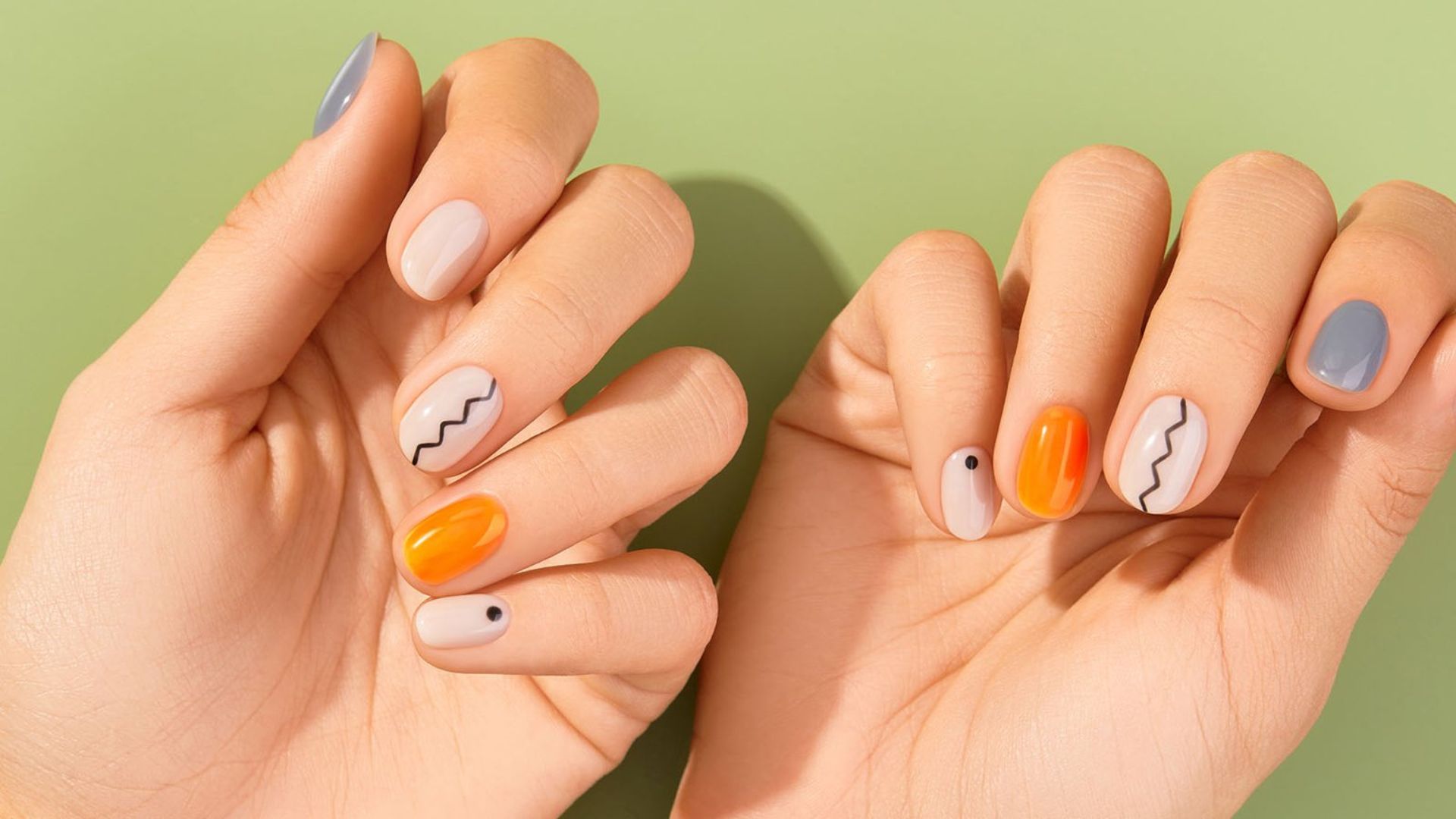 Two hands with creative nail art on a green background. Nails feature zigzag lines, orange polish, and black dots, conveying a playful and modern style.