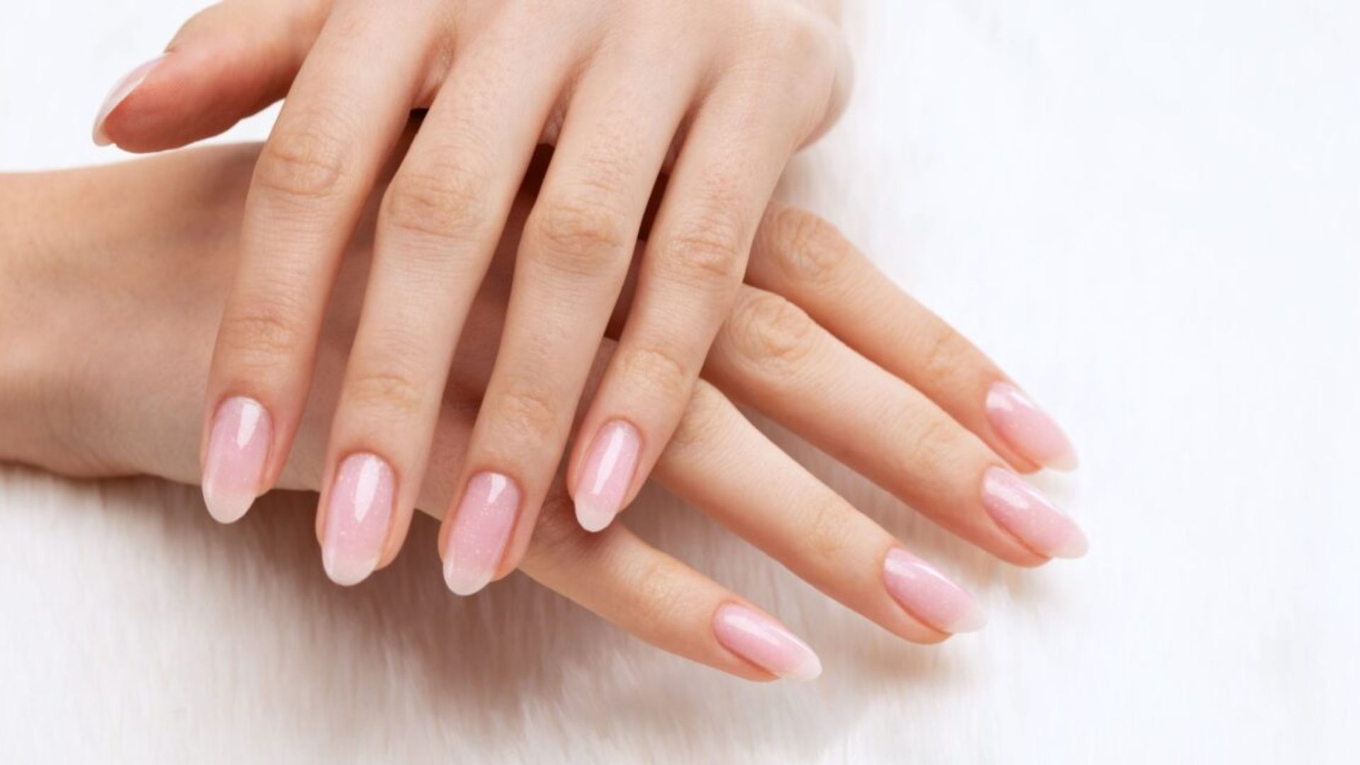 Close-up of hands with neatly manicured nails, featuring a glossy, light pink polish. The background is a soft white, conveying elegance and simplicity.