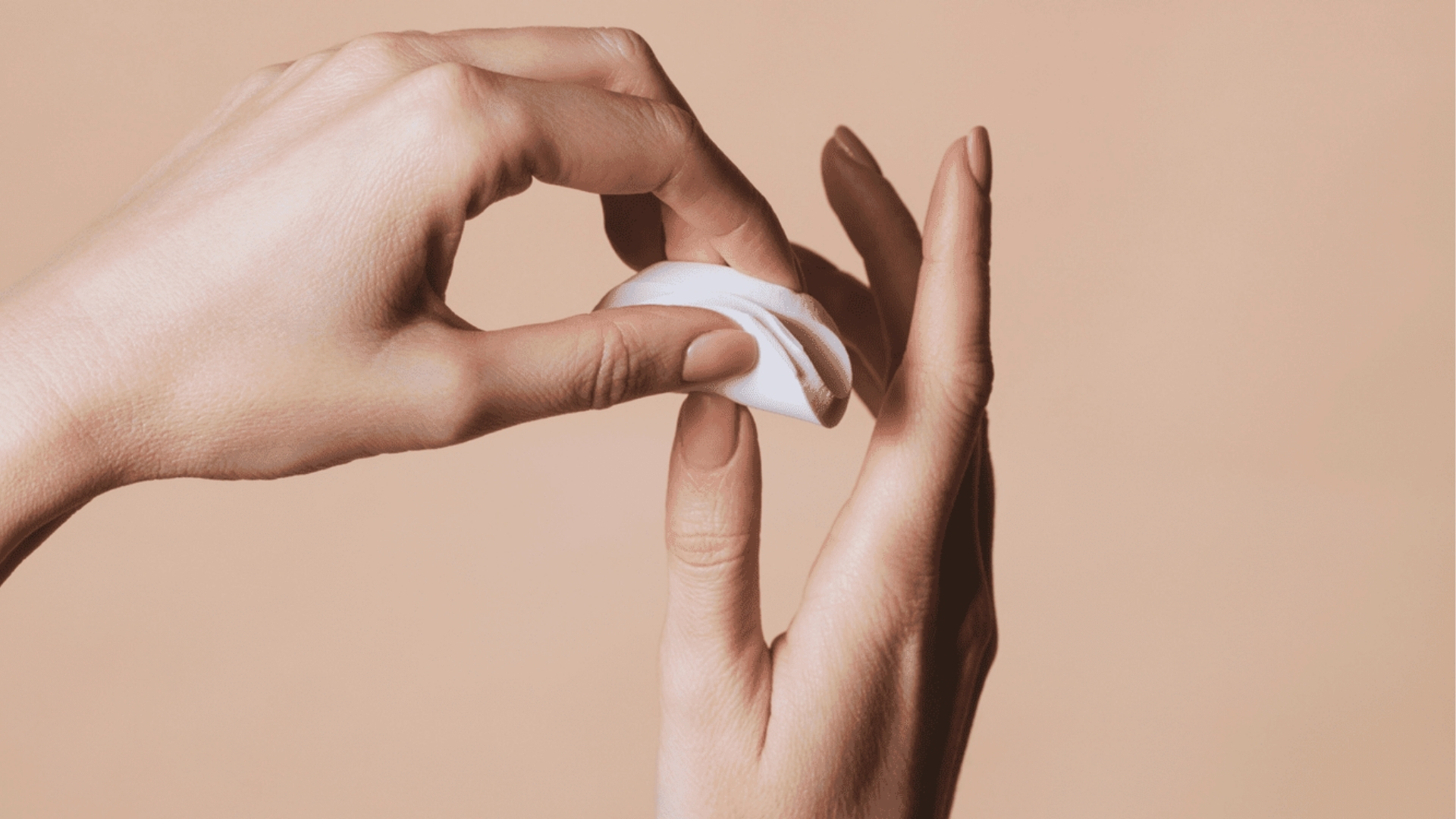 Two hands against a beige background, one holding a cotton pad gently near the fingertips of the other. The tone is soft and serene, suggesting self-care.