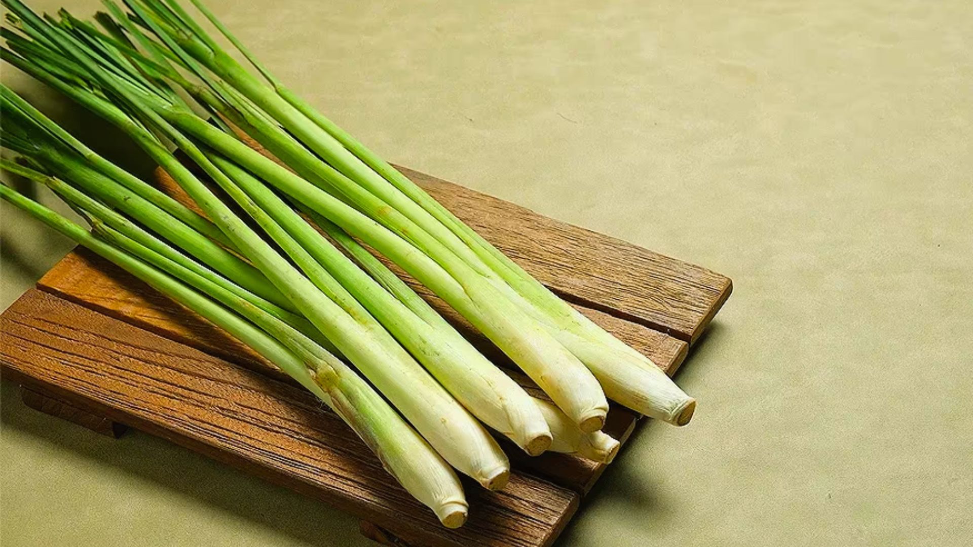 Fresh lemongrass stalks on a wooden cutting board, placed on a beige surface. The vibrant green stalks create a fresh and natural feel.