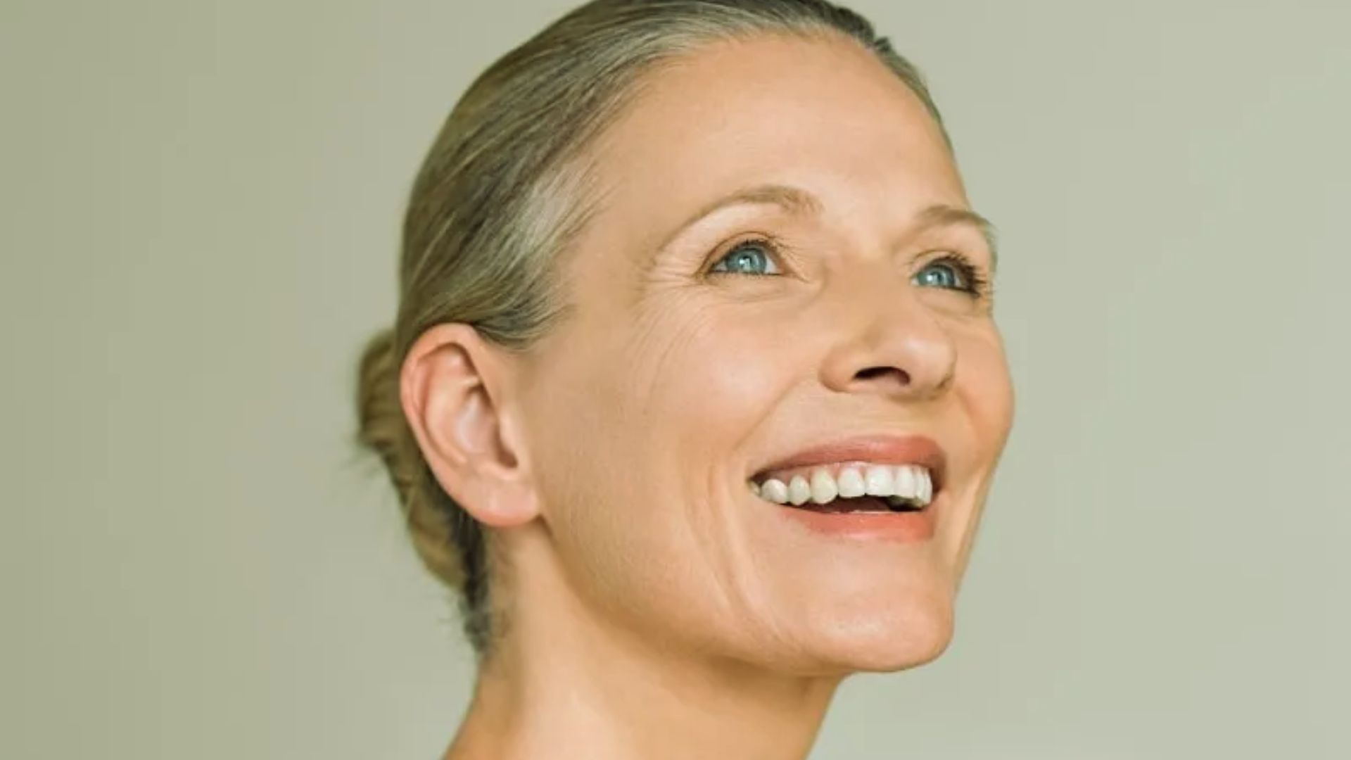 A smiling woman with light hair is gazing upwards against a plain background. Her expression is joyful and serene, conveying a sense of happiness and contentment.