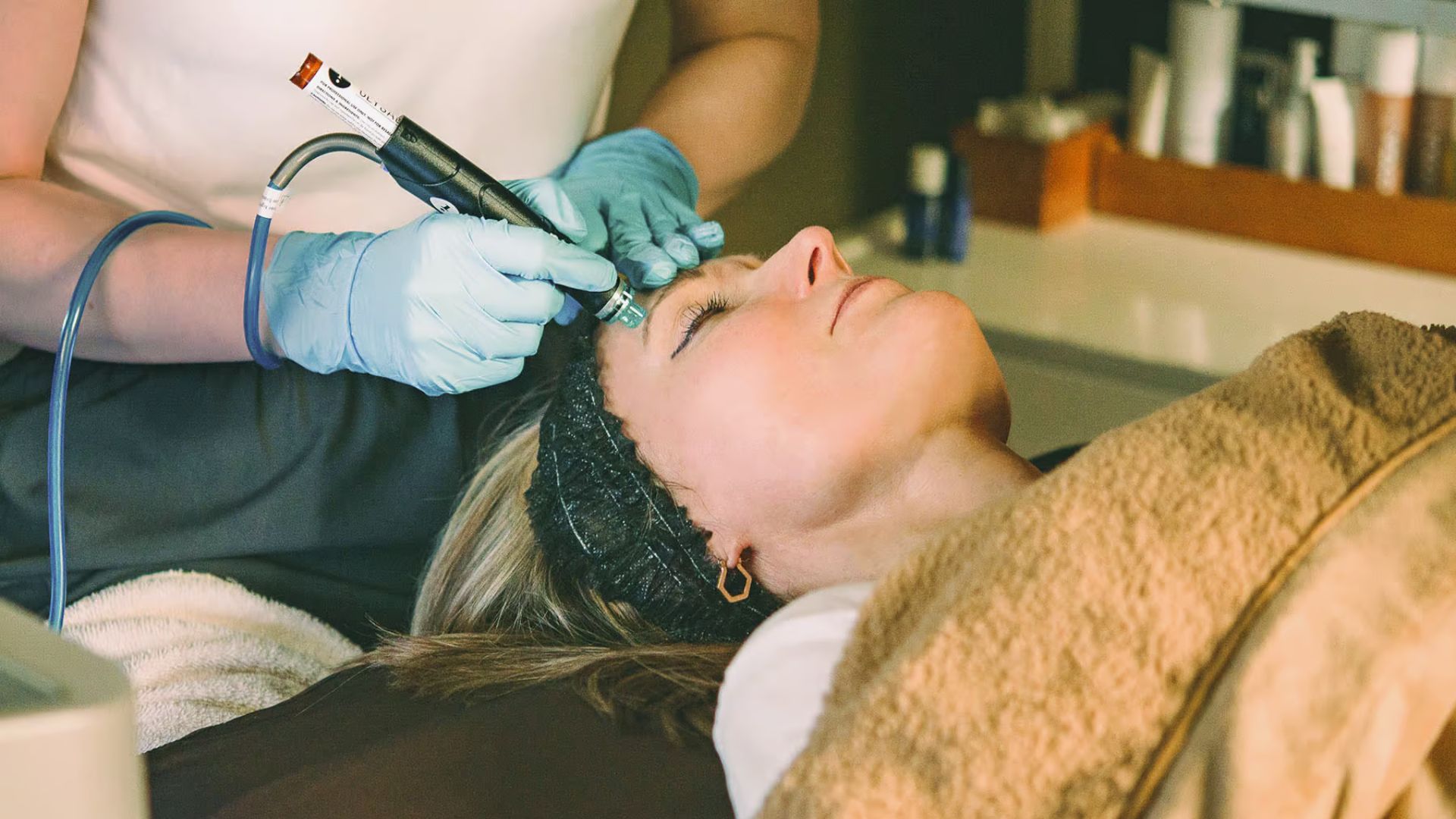 A woman receives a facial treatment with a skincare device in a spa setting, conveying relaxation. She wears a headband; the technician wears gloves.