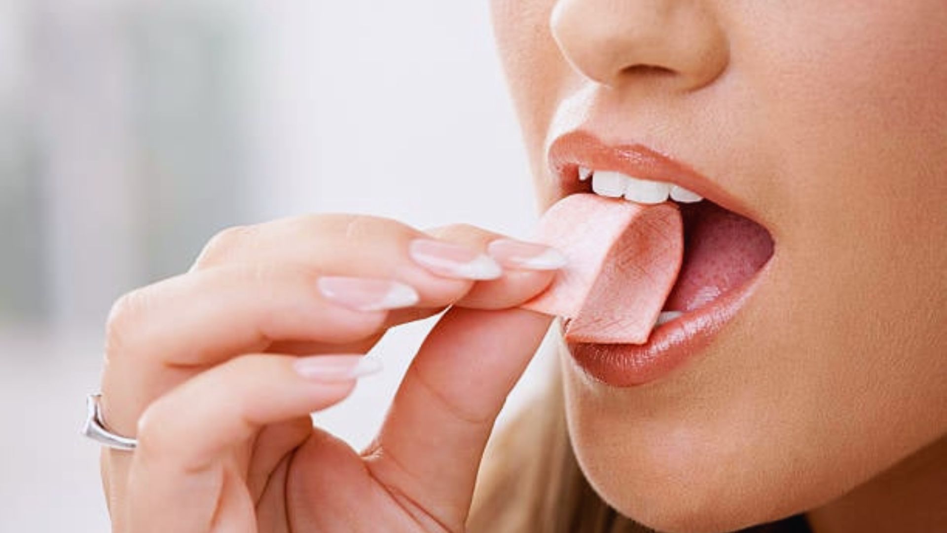 A close-up of a person placing a stick of gum in their mouth. The person has well-manicured nails and natural makeup. The scene conveys freshness.