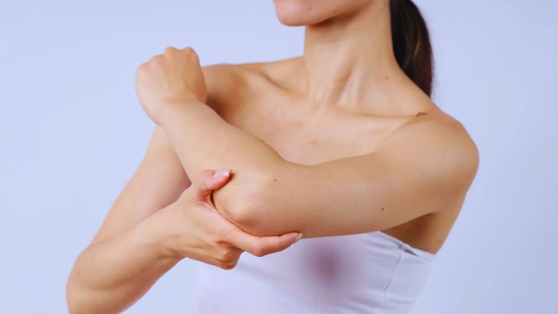 A person gently holds their right elbow with their left hand, wearing a strapless white top. The background is plain, conveying a calm and focused tone.