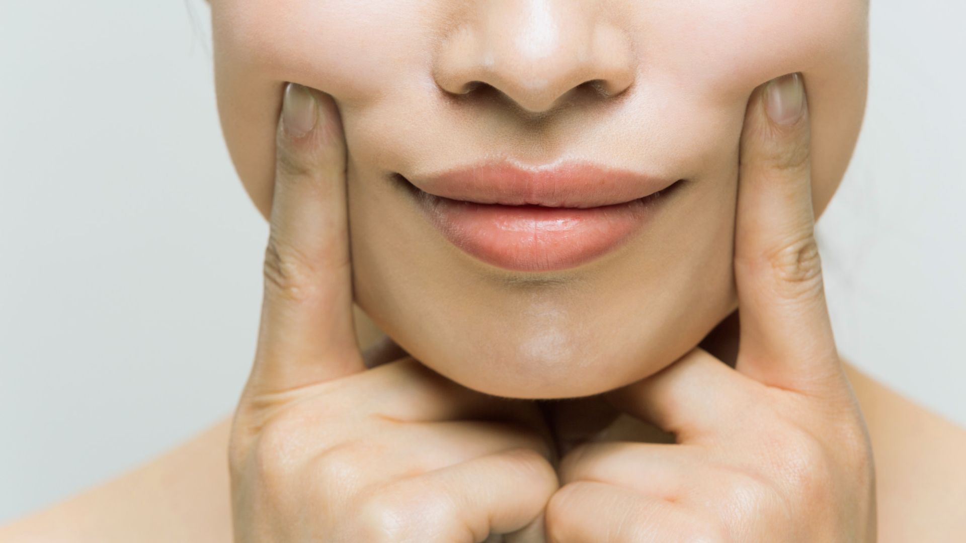 Close-up of a person's lower face with fingers pressing both cheeks. The skin appears smooth, conveying a tone of contentment and relaxation.