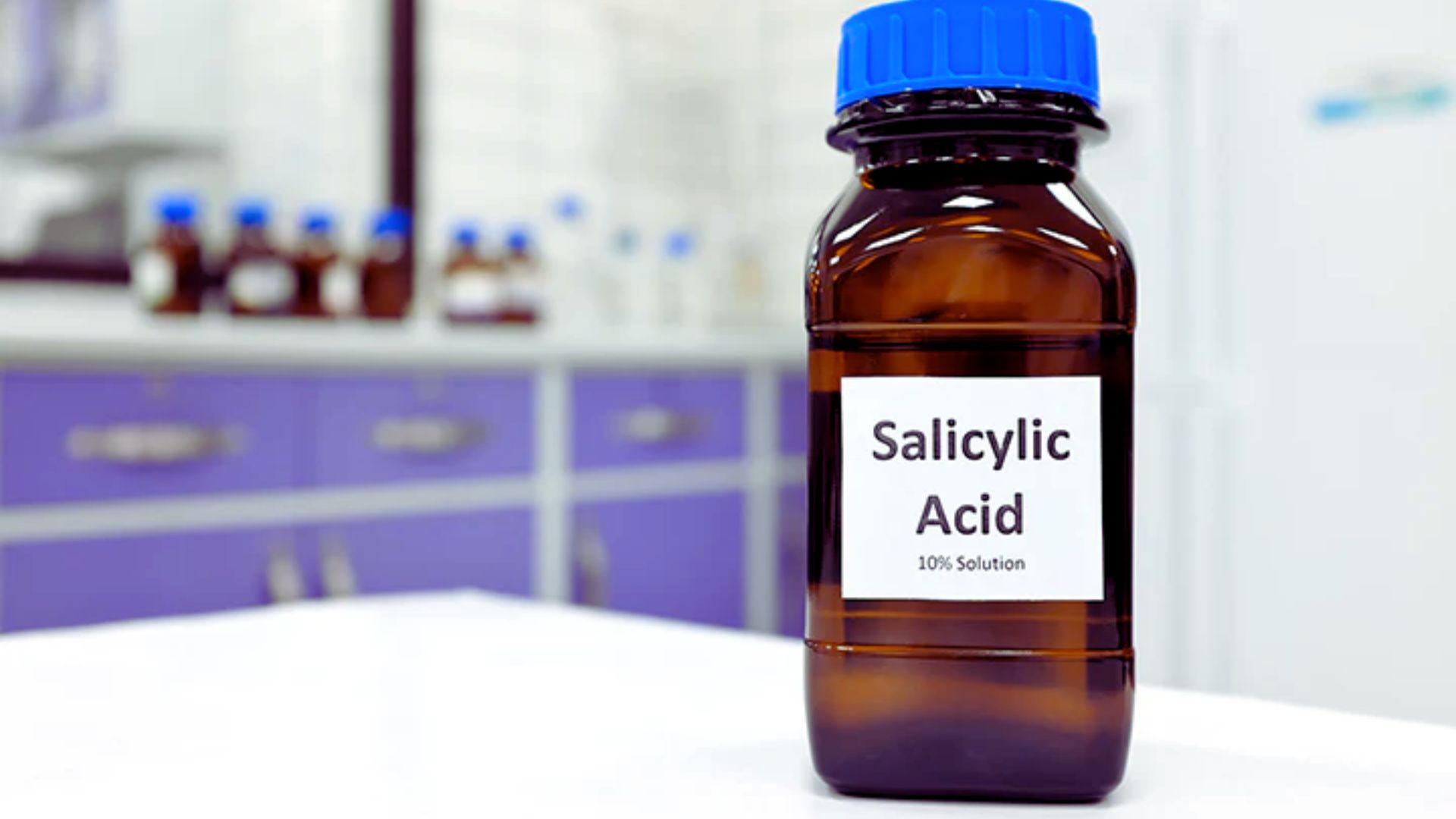 A brown bottle with a blue cap labeled "Salicylic Acid 10% Solution" is on a white countertop in a lab. Purple cabinets are visible in the blurred background.
