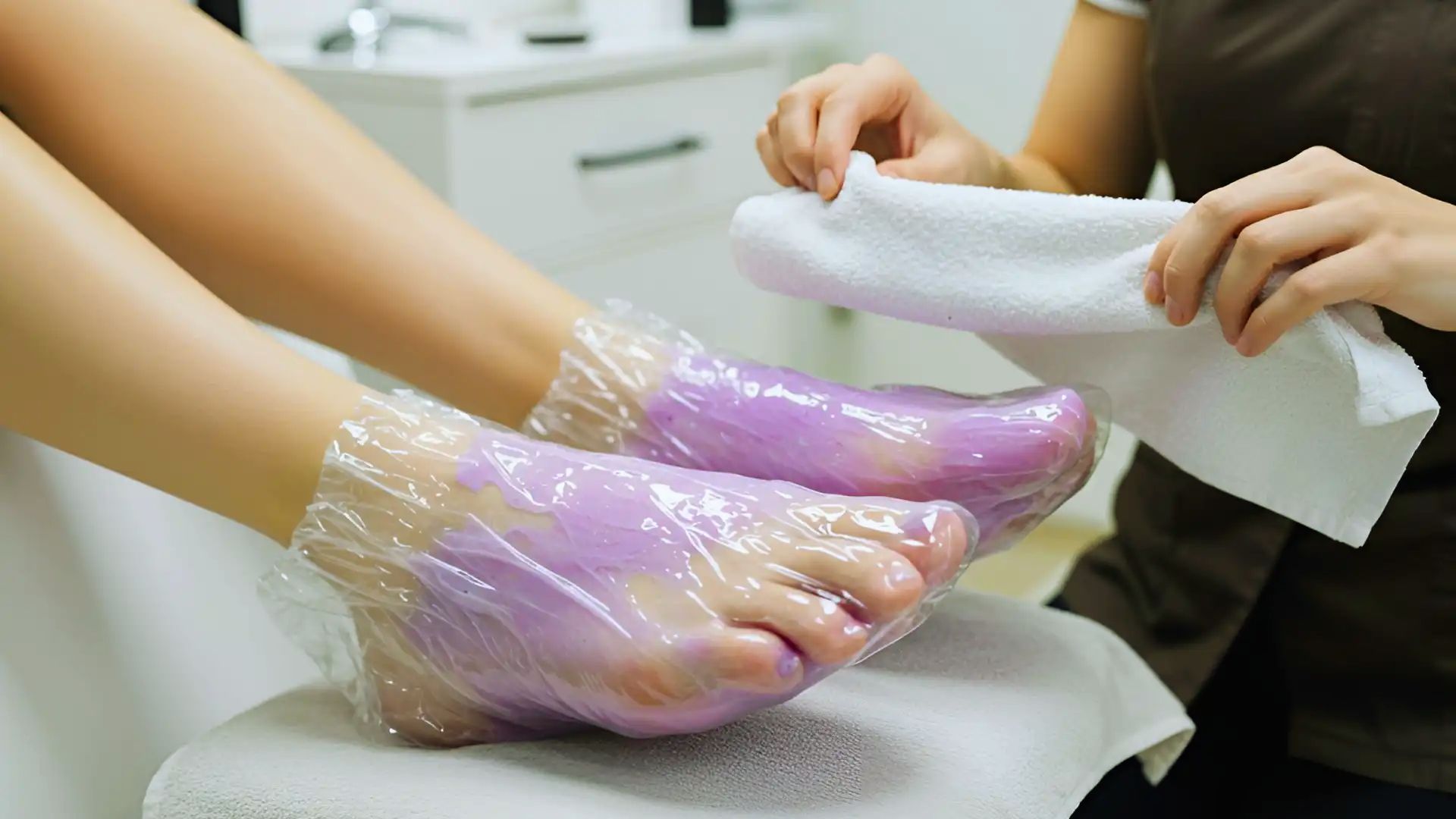 Two feet wrapped in transparent plastic with a purple gel, receiving a pedicure. A person holds a white towel, suggesting care and relaxation.