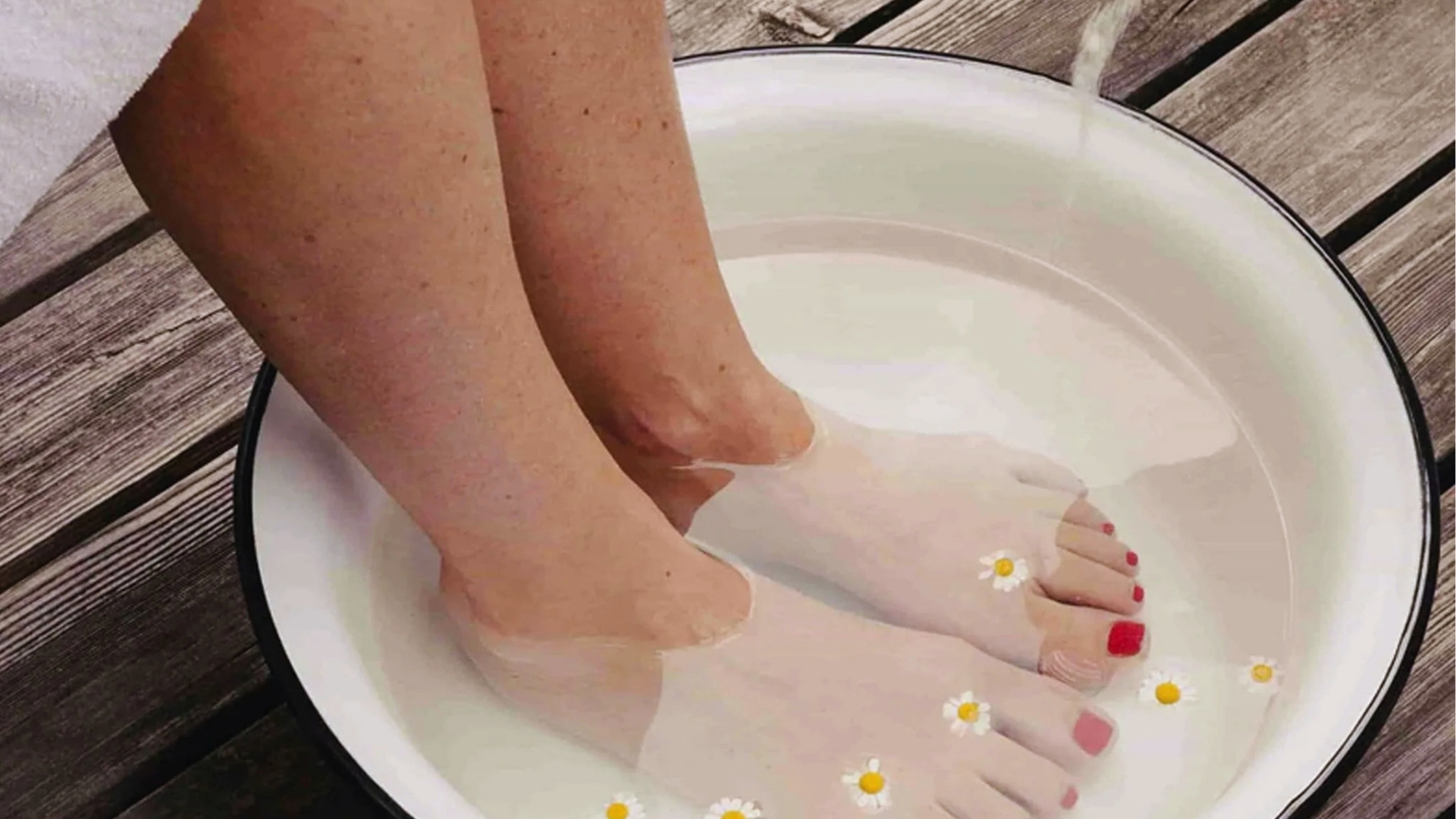 Close-up of feet soaking in a white basin of water with floating daisies on a wooden deck. The scene conveys relaxation and a spa-like atmosphere.