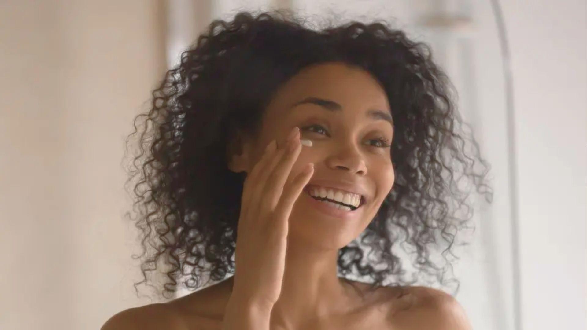 Smiling woman with curly hair applies skincare product under her eye during a recovery night.