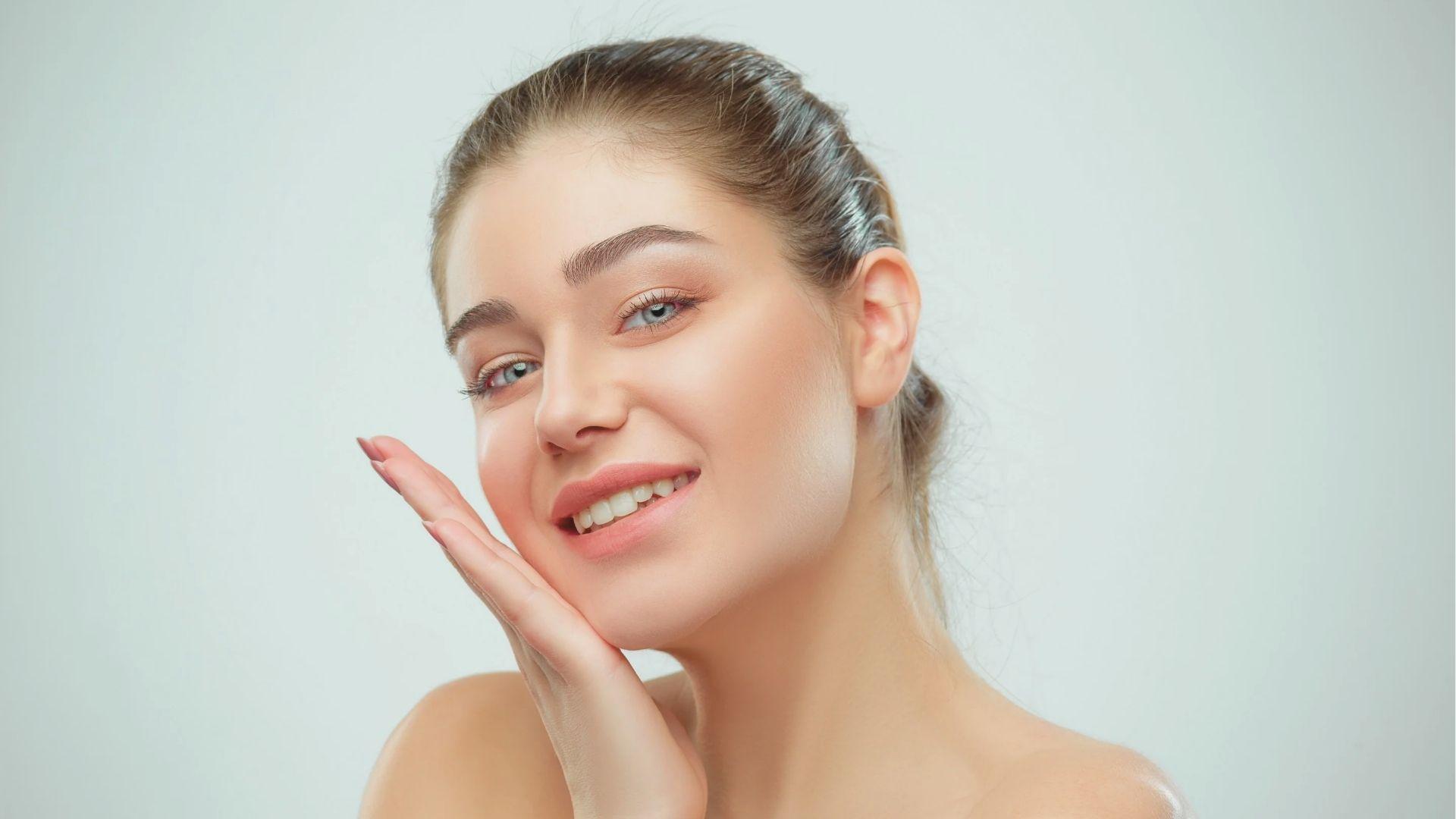 A smiling woman with tied-back hair and flawless skin gently framing her face with one hand.