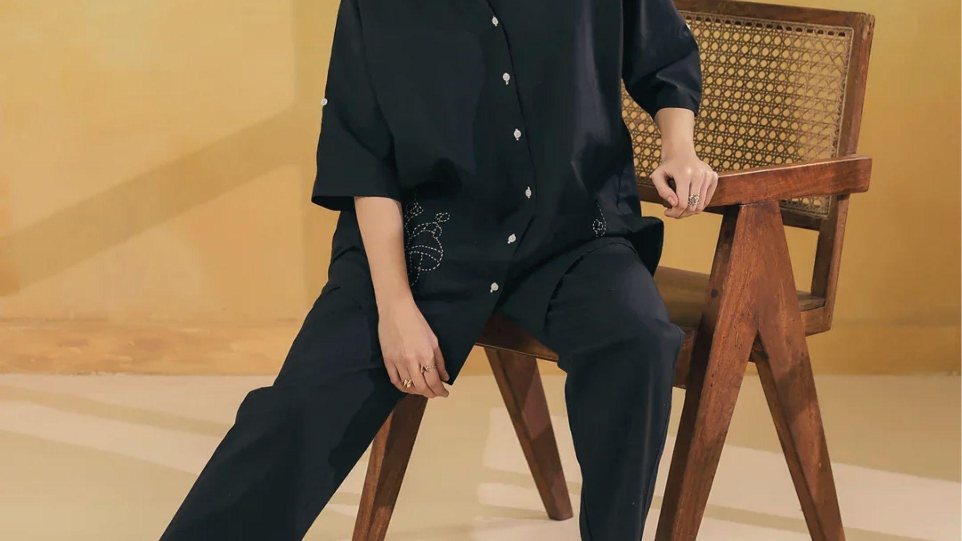 Person in oversized black button shirt and wide pants sits on chair, loose fit prevents spray tan lines