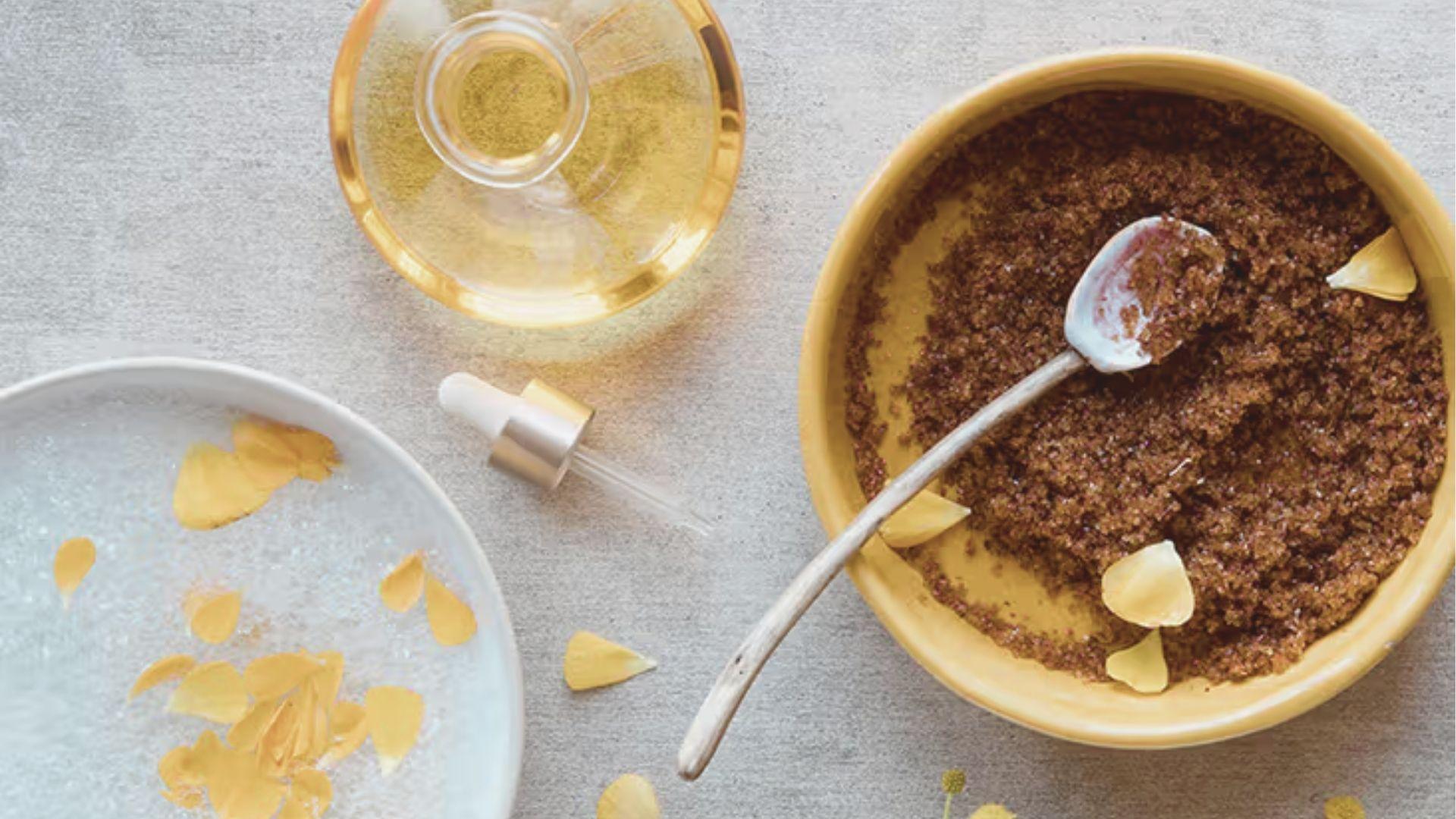 A yellow bowl of brown sugar scrub with a spoon, alongside oil and rose petals for a homemade treatment.