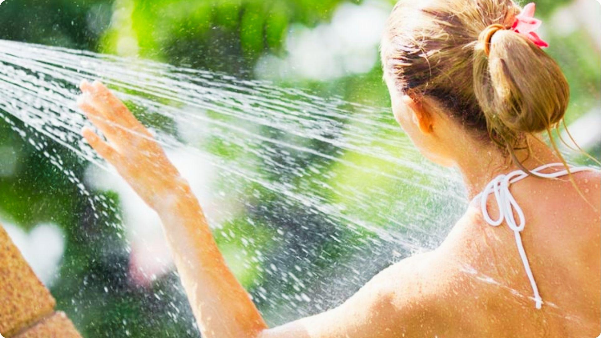 A woman stands under an outdoor shower spray with greenery in the background.
