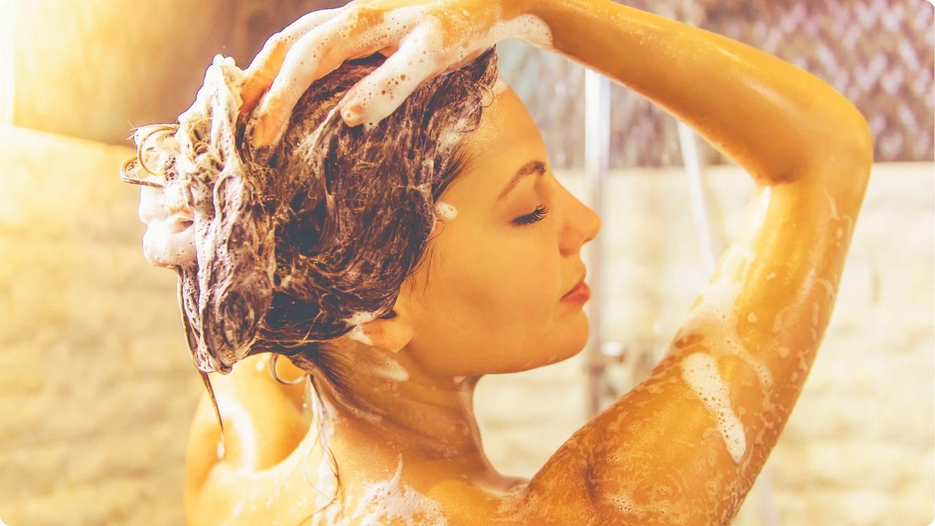 A woman washes her hair in the shower with shampoo suds and warm lighting.