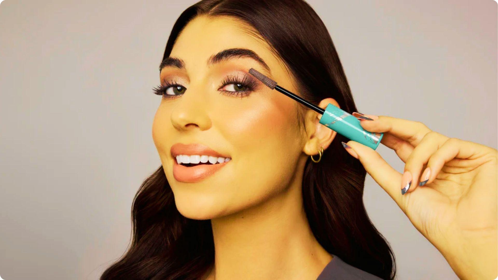 Smiling person holding a turquoise mascara tube, applying the wand to curled lashes.