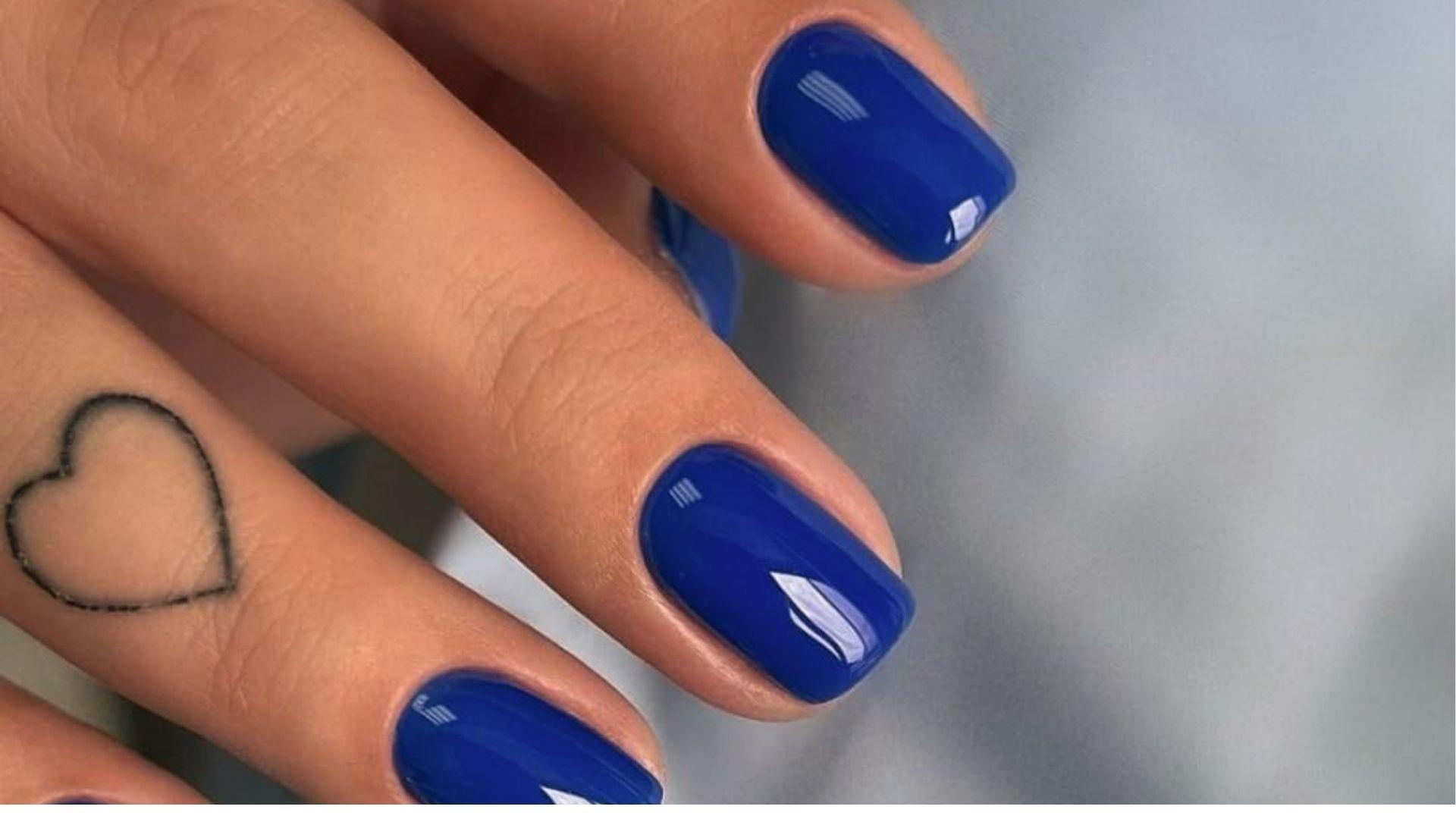 Short square dip nails in solid glossy cobalt blue on hand with heart tattoo on ring finger.