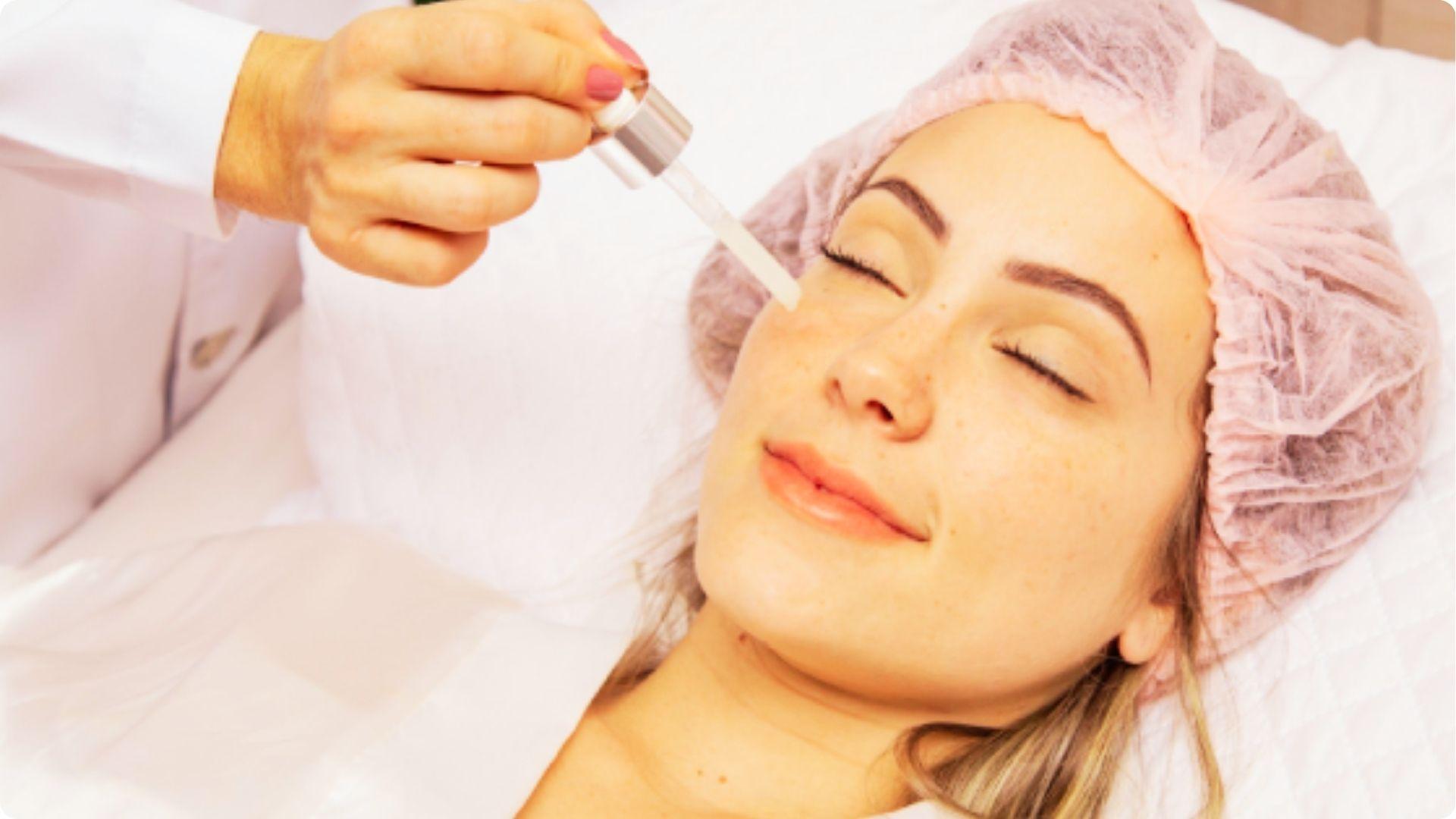 Esthetician applies hyaluronic acid serum to a relaxed client’s face during a facial treatment in a clinic.