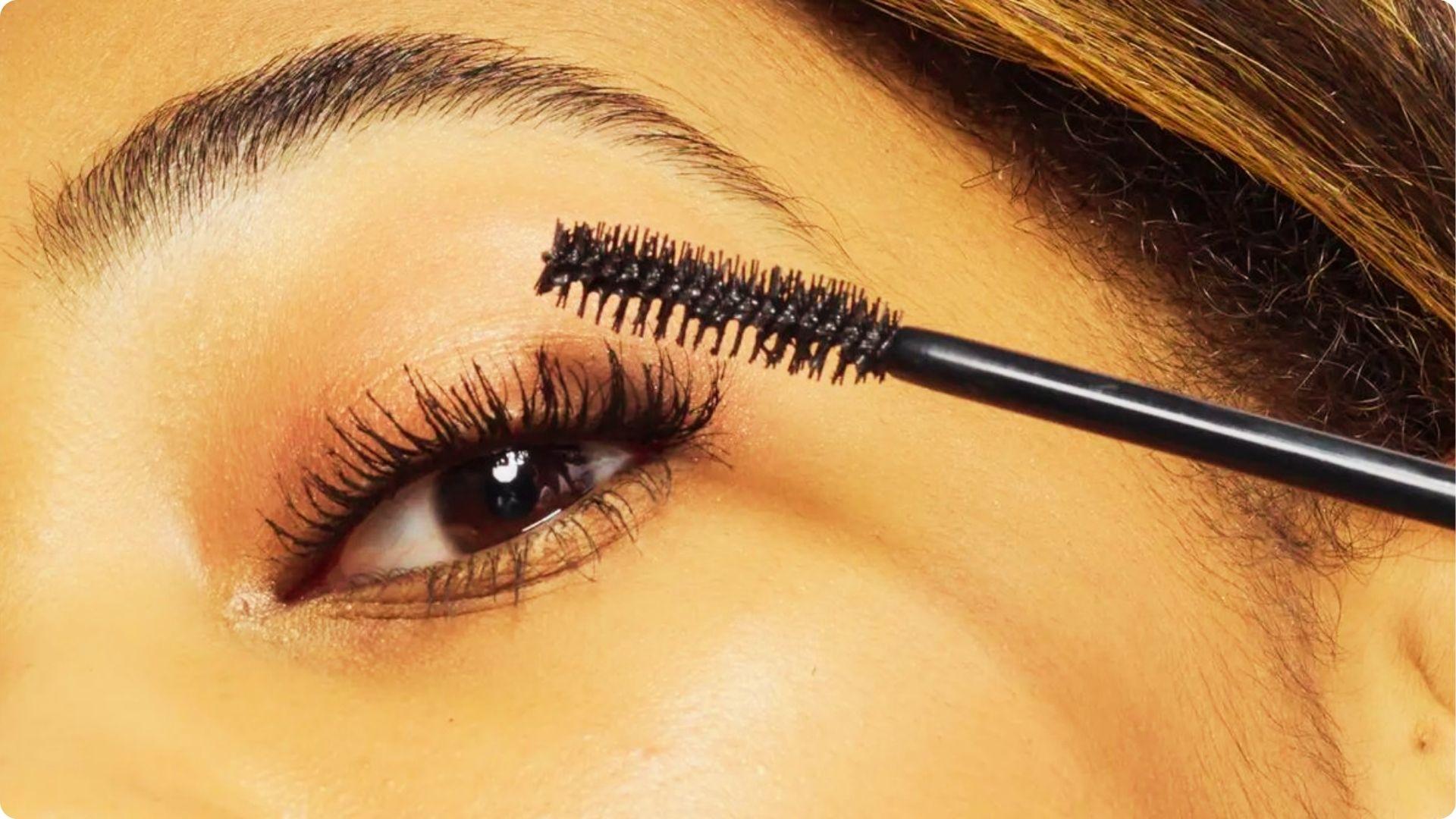 Macro shot of a mascara wand coating long, curled lashes on a brown eye.