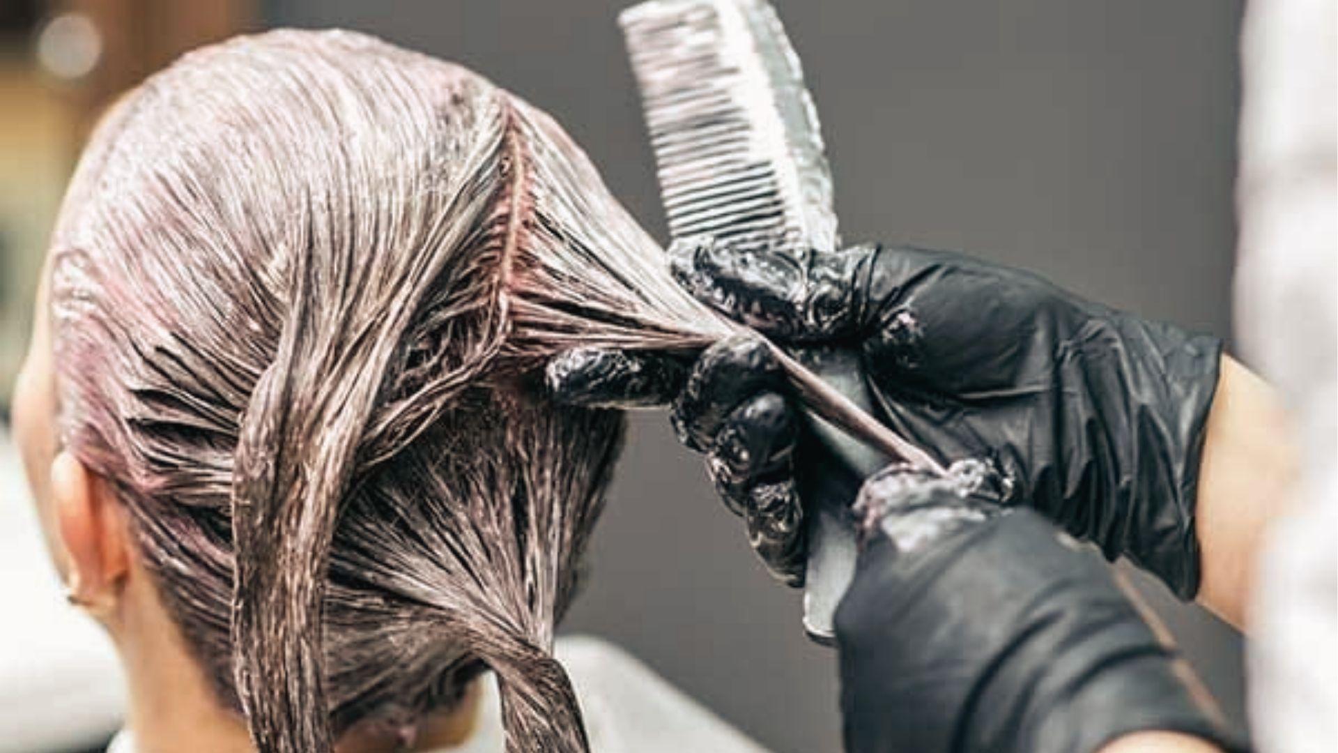 Gloved stylist combing bleach through wet hair roots, ensuring full saturation during the timed process.