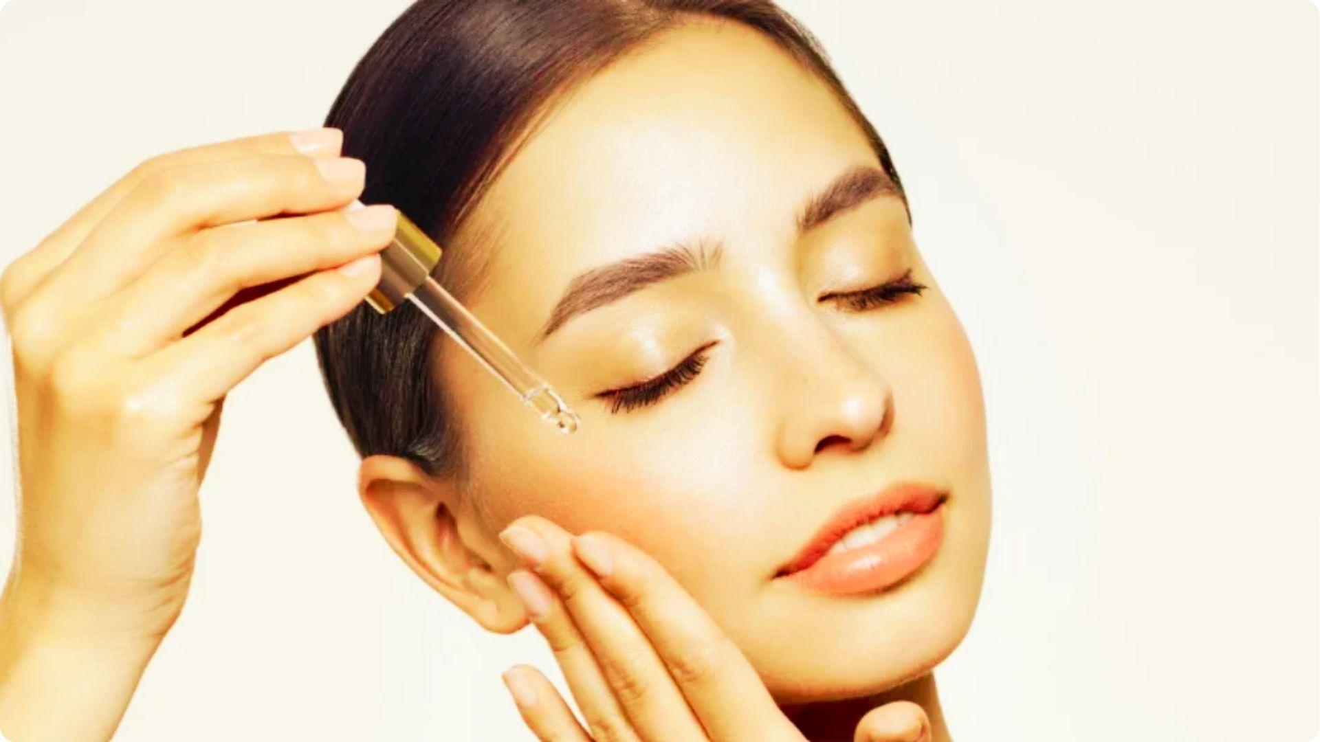 Woman with eyes closed gently applies hyaluronic acid serum to her cheek using a gold-topped dropper.