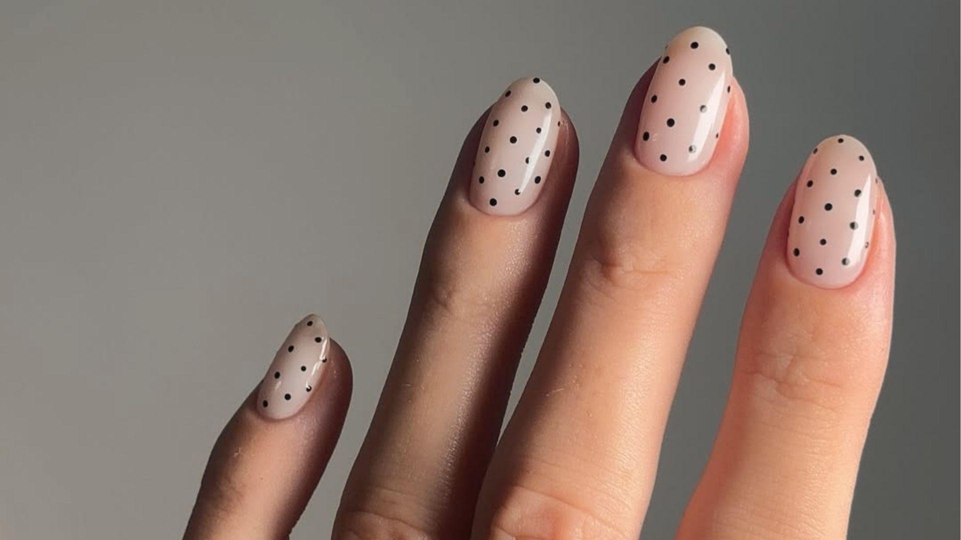 Short oval dip nails in glossy milky nude base with tiny scattered black polka dots for retro vibe.