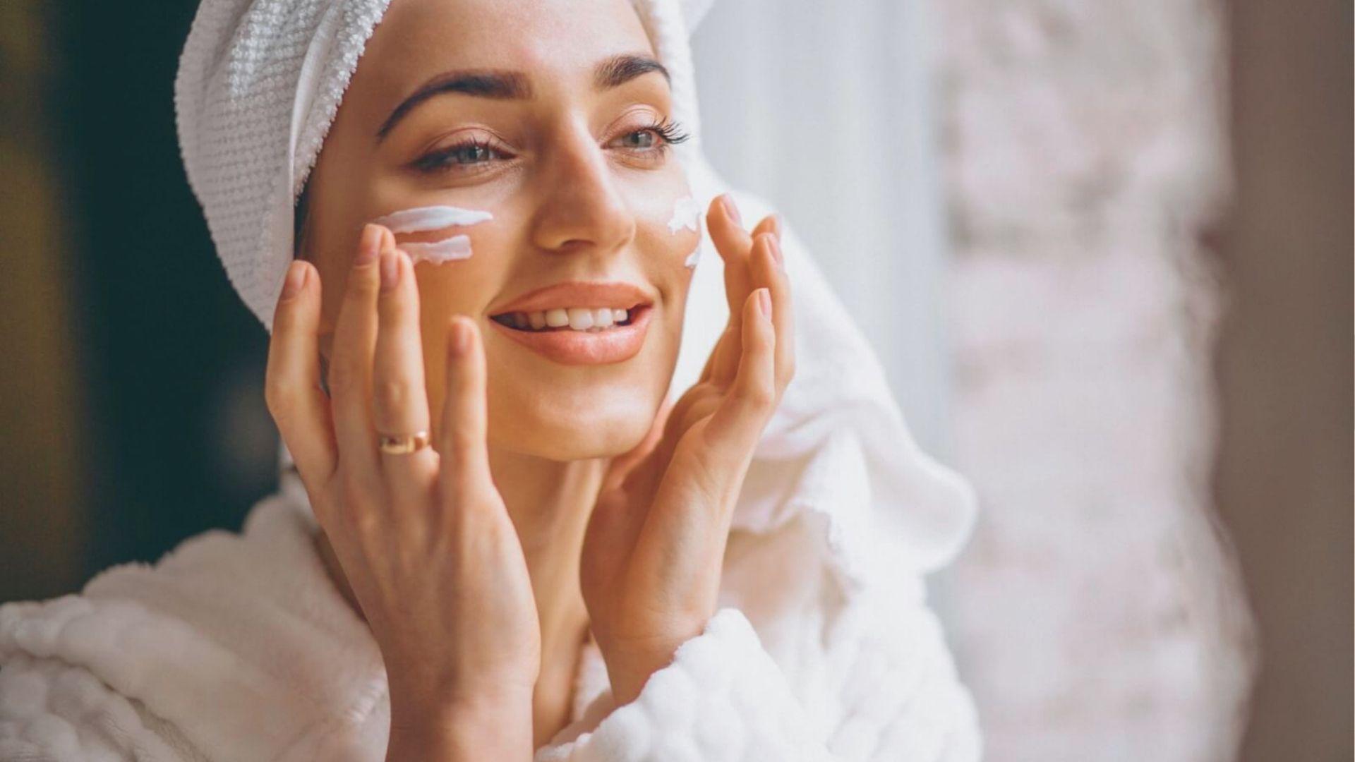 Woman in a robe and towel smiles while applying cream to her face during a recovery step.