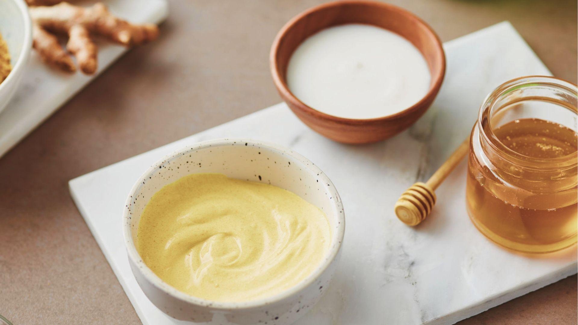 Homemade honey face mask in a speckled bowl next to honey, yogurt, and fresh turmeric root.