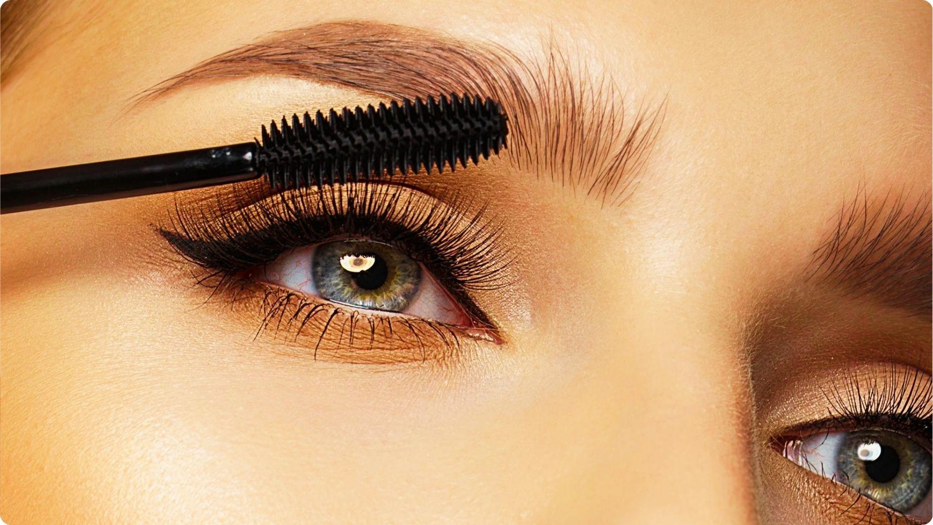 Extreme close-up of a mascara wand coating top lashes and brows of a person with light eyes.