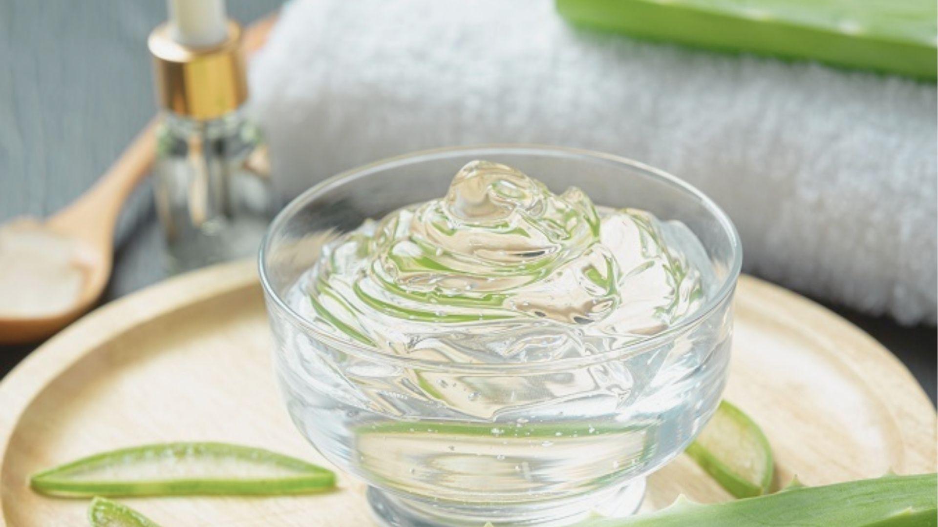 A glass bowl of fresh aloe vera gel with aloe leaves, a dropper bottle, and a white towel nearby.