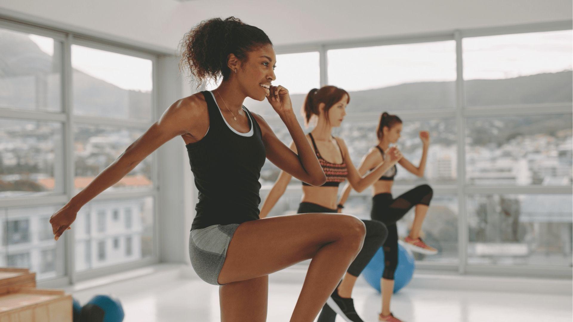 Three women doing cardio in a bright studio with large windows overlooking a city.