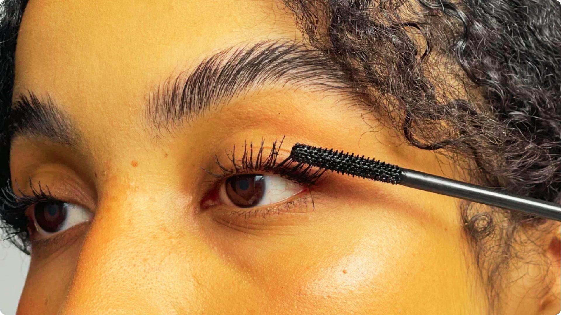 Close-up of a person with curly hair applying black mascara to upper lashes with a textured wand.