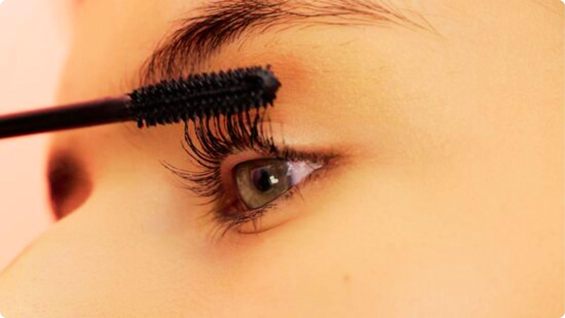 Close-up of a mascara brush separating dark lashes on a green eye with winged eyeliner.